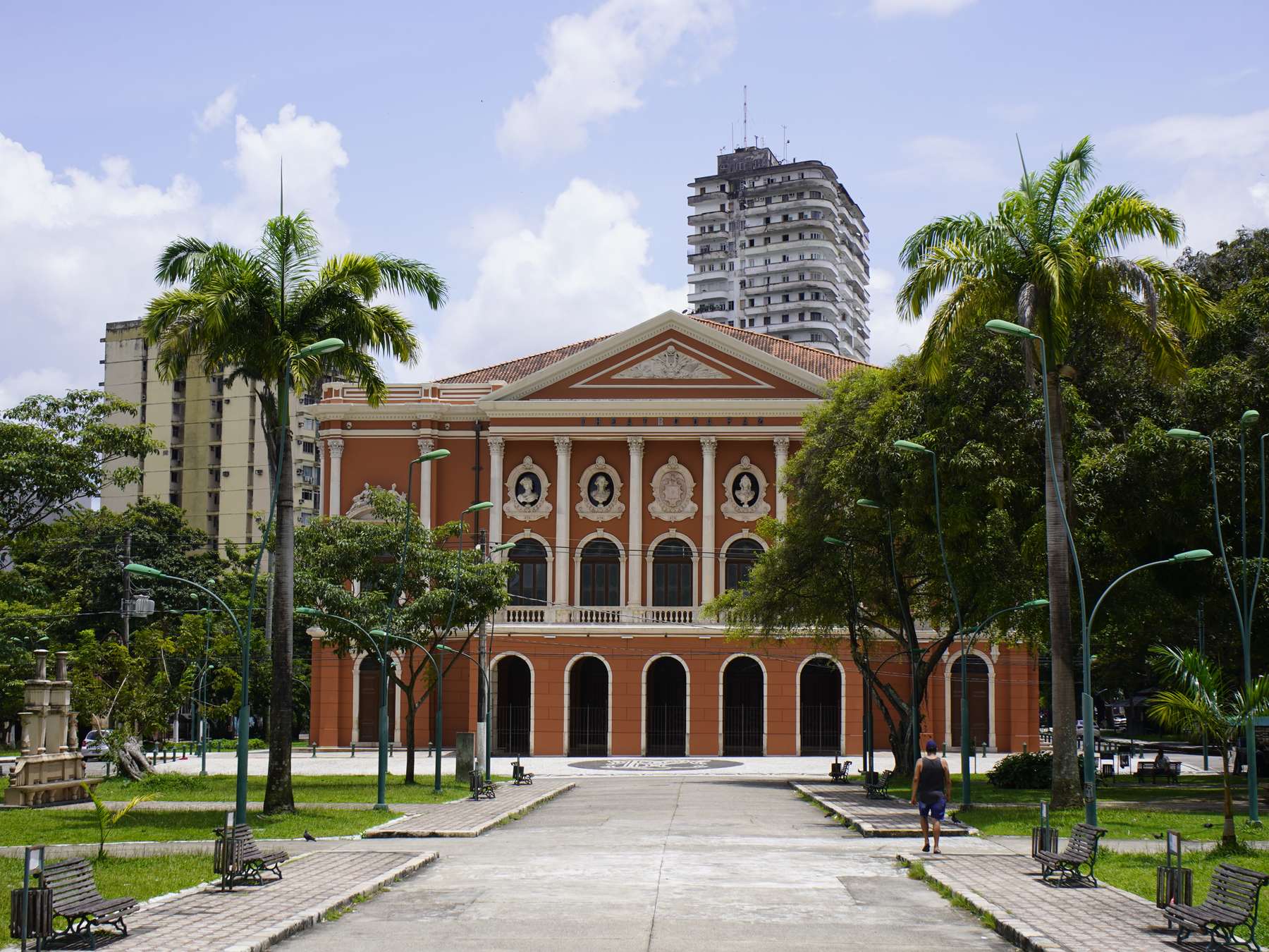 Fachada neoclássica do Theatro da Paz vista da Praça da República, com palmeiras, bancos e o calçadão em primeiro plano, sob céu azul com nuvens