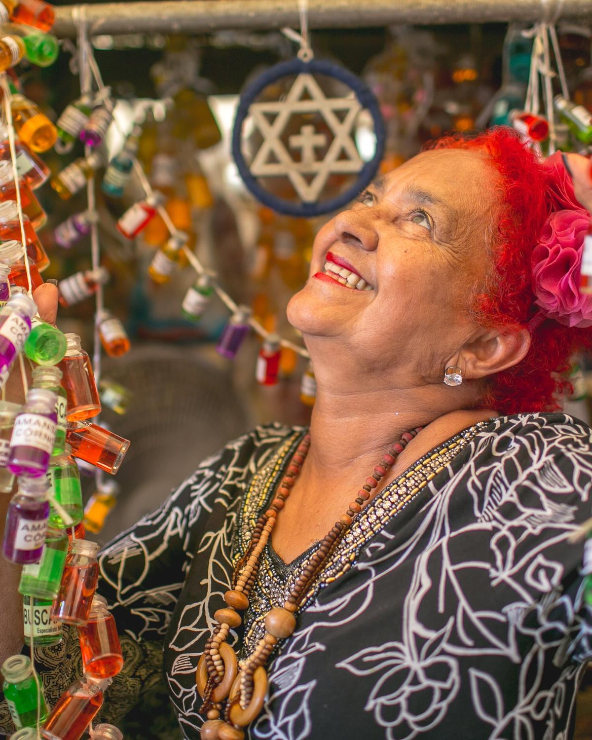 Erveira sorrindo na banca do Ver-o-Peso, cercada por frascos coloridos de perfumes e banhos de ervas pendurados.