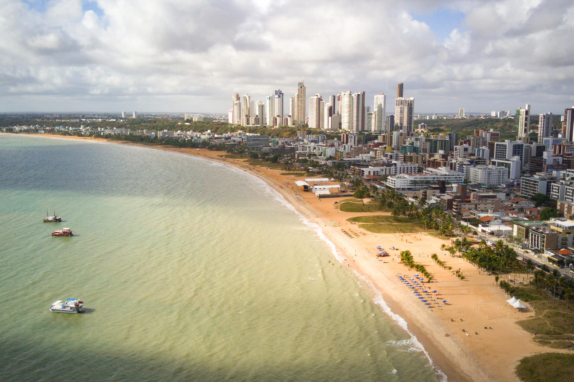 Vista aérea da orla de João Pessoa com a praia de Tambaú, a faixa de prédios da cidade e barcos de passeio ancorados no mar esverdeado