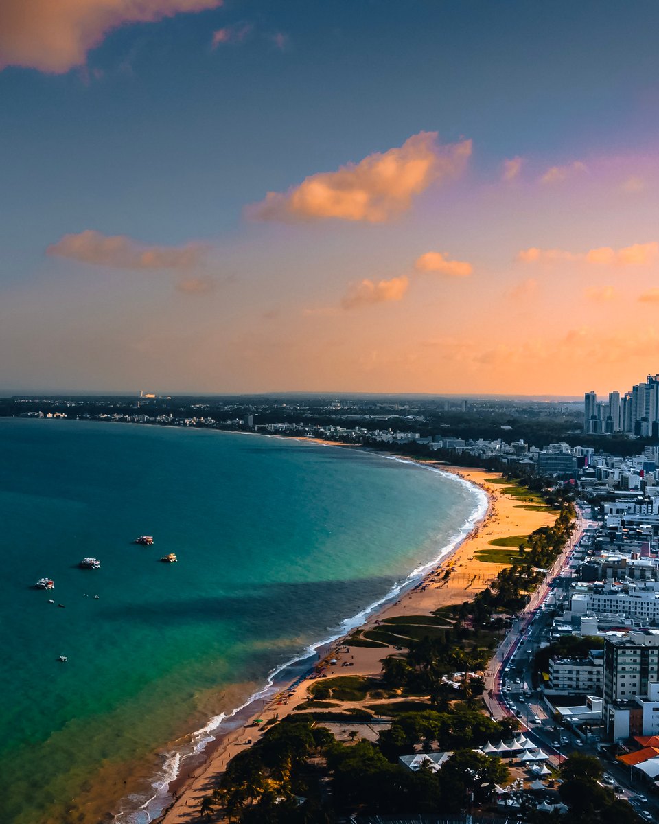 Vista aérea da orla de João Pessoa no fim da tarde, com a curva da praia, barcos ancorados no mar esverdeado, a avenida da beira-mar e a faixa de prédios da cidade ao fundo