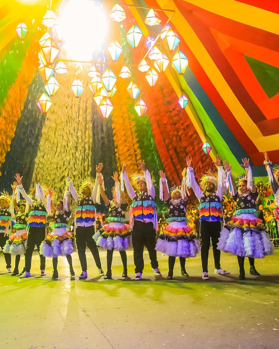 Casais de quadrilha junina com os braços erguidos no Parque do Povo, em Campina Grande, sob a cobertura colorida de bandeirinhas e balões iluminados