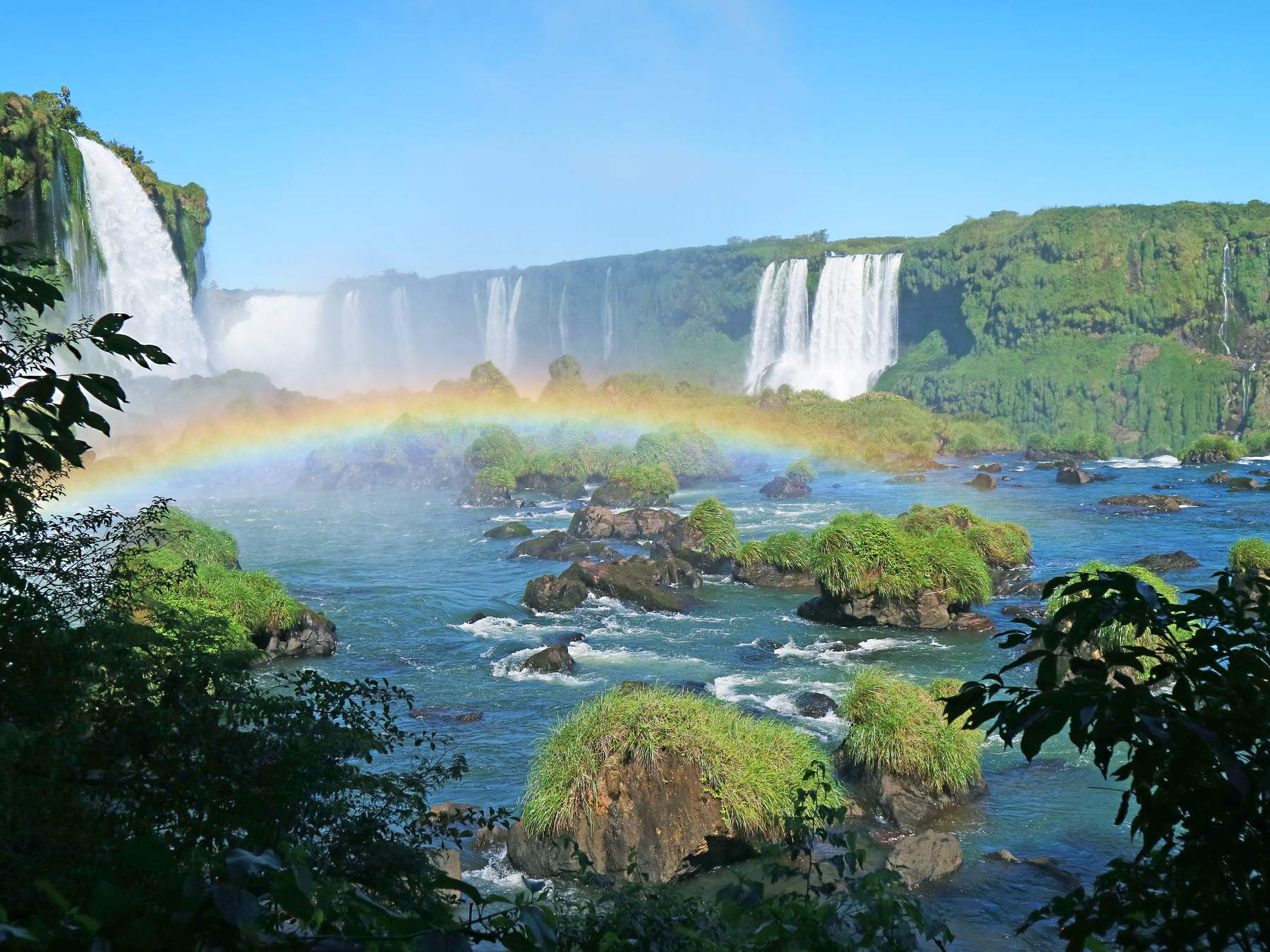 Cataratas do Iguaçu vistas do lado brasileiro, com várias quedas ao fundo, um arco-íris sobre o rio e ilhas de vegetação entre as corredeiras.