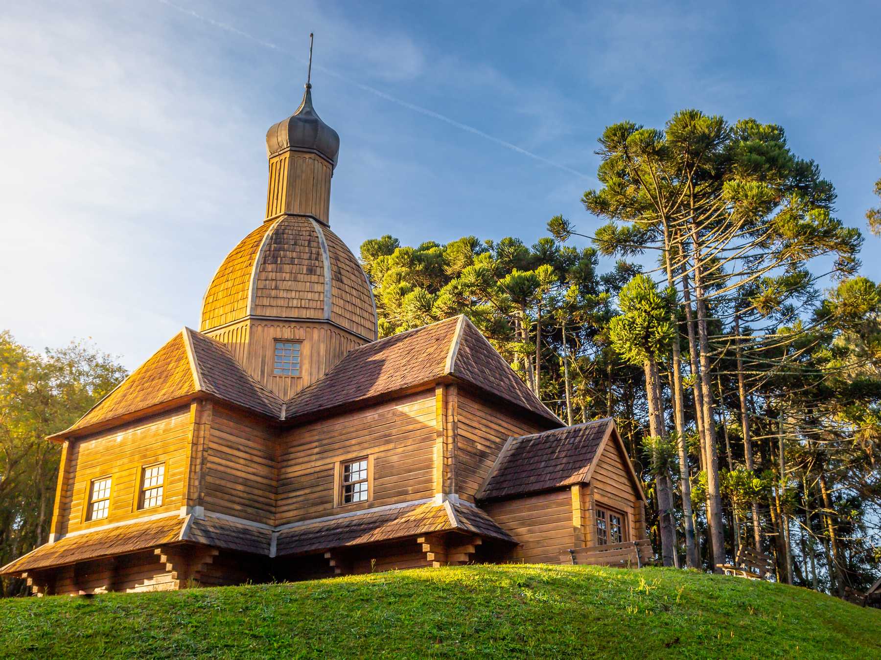 Igreja ucraniana de madeira com cúpula dourada no Memorial Ucraniano, cercada de araucárias, sob a luz do fim de tarde.
