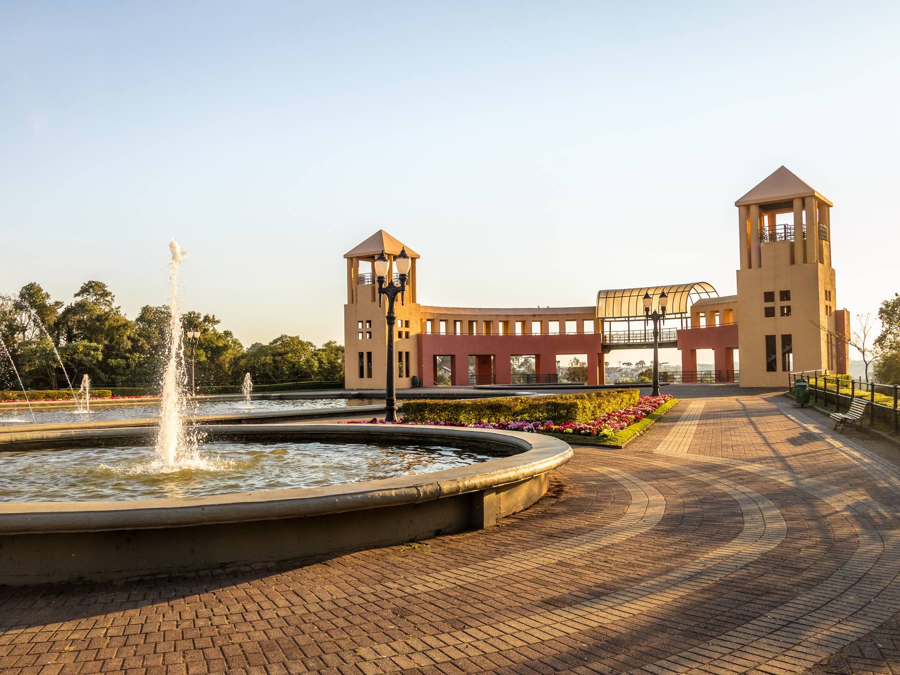 Chafariz e o portal com duas torres do Parque Tanguá, com piso de tijolos iluminado pelo sol baixo.