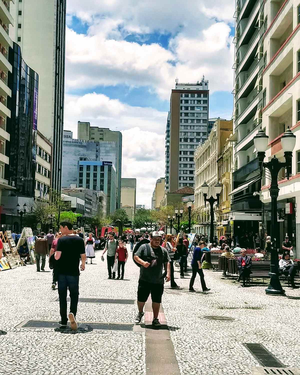 Pedestres caminhando pelo calçadão da Rua XV de Novembro, no centro de Curitiba, entre prédios altos, postes antigos e lojas.