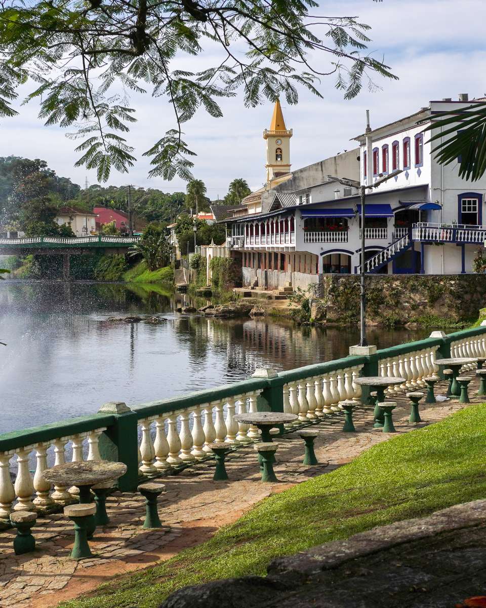 Margem do rio Nhundiaquara no centro histórico de Morretes, com balaustrada, mesas de pedra no gramado, um chafariz no rio, casarões coloniais e a torre da igreja ao fundo.