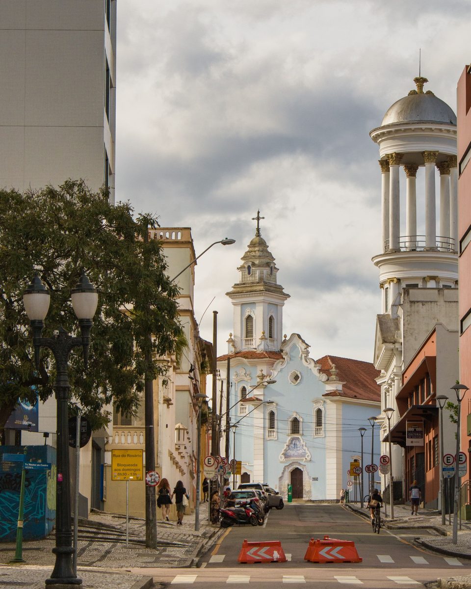 Rua de pedestres e ciclistas no Largo da Ordem, centro histórico de Curitiba, com a Igreja do Rosário azul e branca ao fundo, a torre do Belvedere à direita e postes antigos.