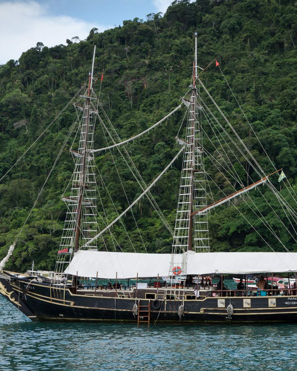 Escuna de madeira de dois mastros navegando na água verde da baía de Paraty, com turistas no convés coberto e a encosta de mata atlântica ao fundo.