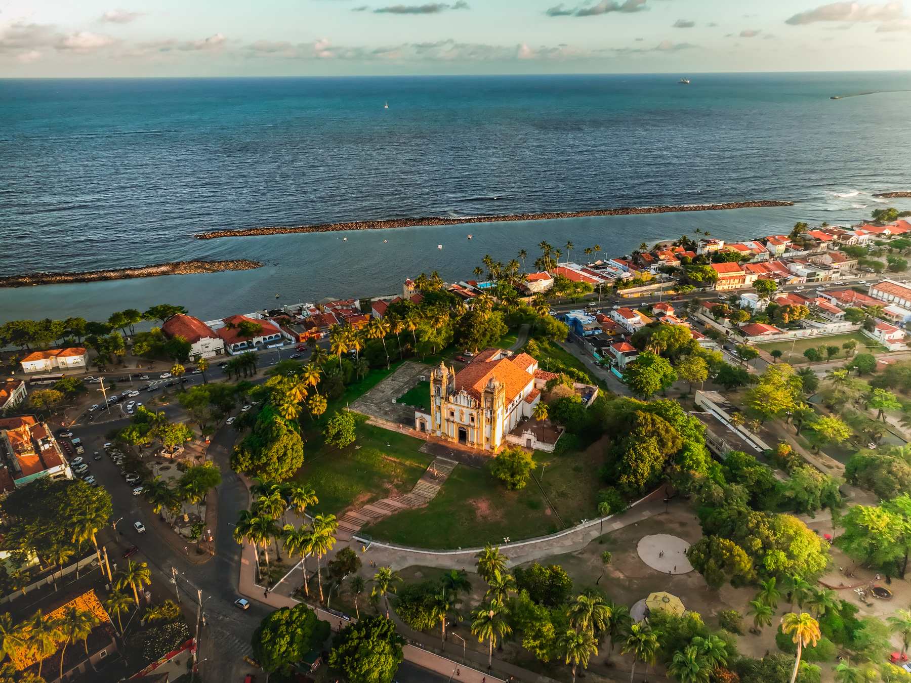 Igreja do Carmo, em Olinda, vista do alto no fim da tarde, cercada de coqueiros e gramado, com o casario da orla, os quebra-mares e o mar ao fundo.