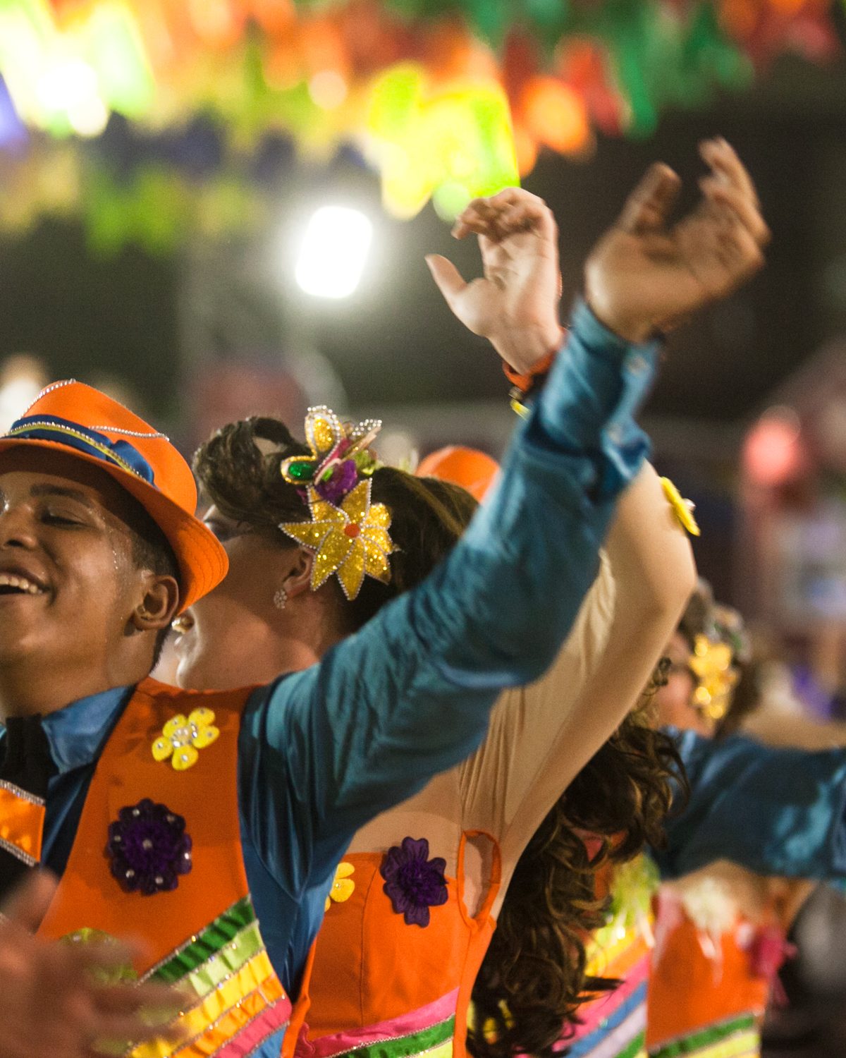 Dançarino de quadrilha junina com chapéu laranja e colete bordado de flores sorrindo de braços erguidos, com outros brincantes e bandeirinhas atrás, à noite no São João de Caruaru.