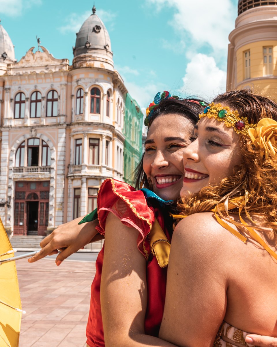 Duas passistas de frevo abraçadas e sorrindo, com fantasias coloridas, flores no cabelo e sombrinhas amarela e rosa, numa praça do Bairro do Recife com casarões ao fundo.