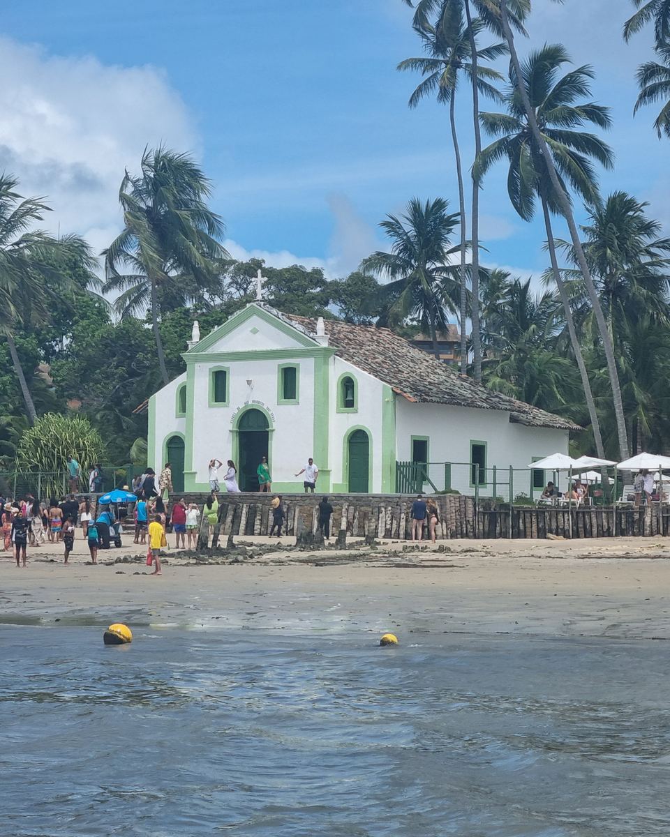 Capela de São Benedito, branca com detalhes verdes, entre coqueiros na Praia dos Carneiros, com banhistas na areia, guarda-sóis e o mar raso em primeiro plano.