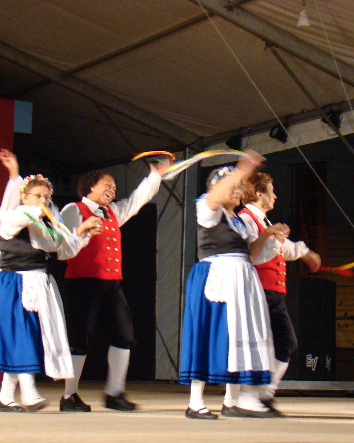 Grupo de mulheres com trajes típicos alemães, coletes vermelhos, saias azuis e aventais brancos, dançando e rindo com fitas coloridas no palco da Bauernfest.