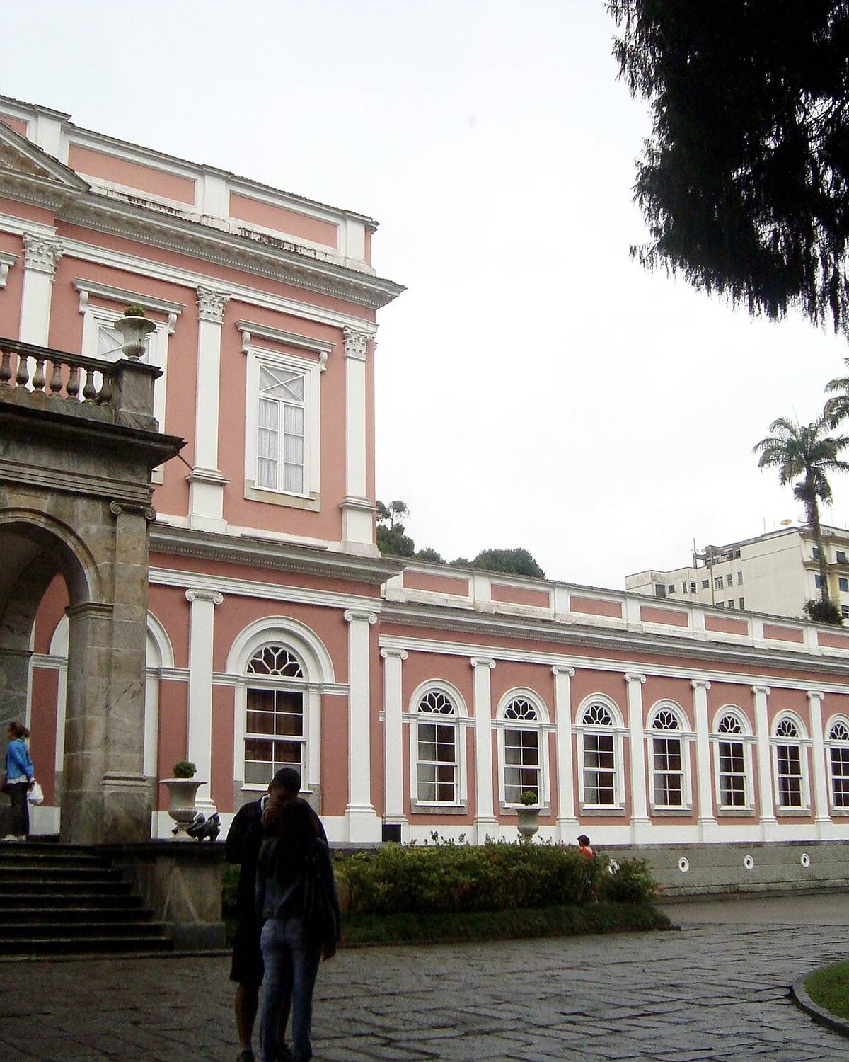 Turistas na entrada do Museu Imperial, em Petrópolis, com o palácio rosa de janelas brancas, palmeiras ao fundo e um visitante mexendo na câmera em primeiro plano.