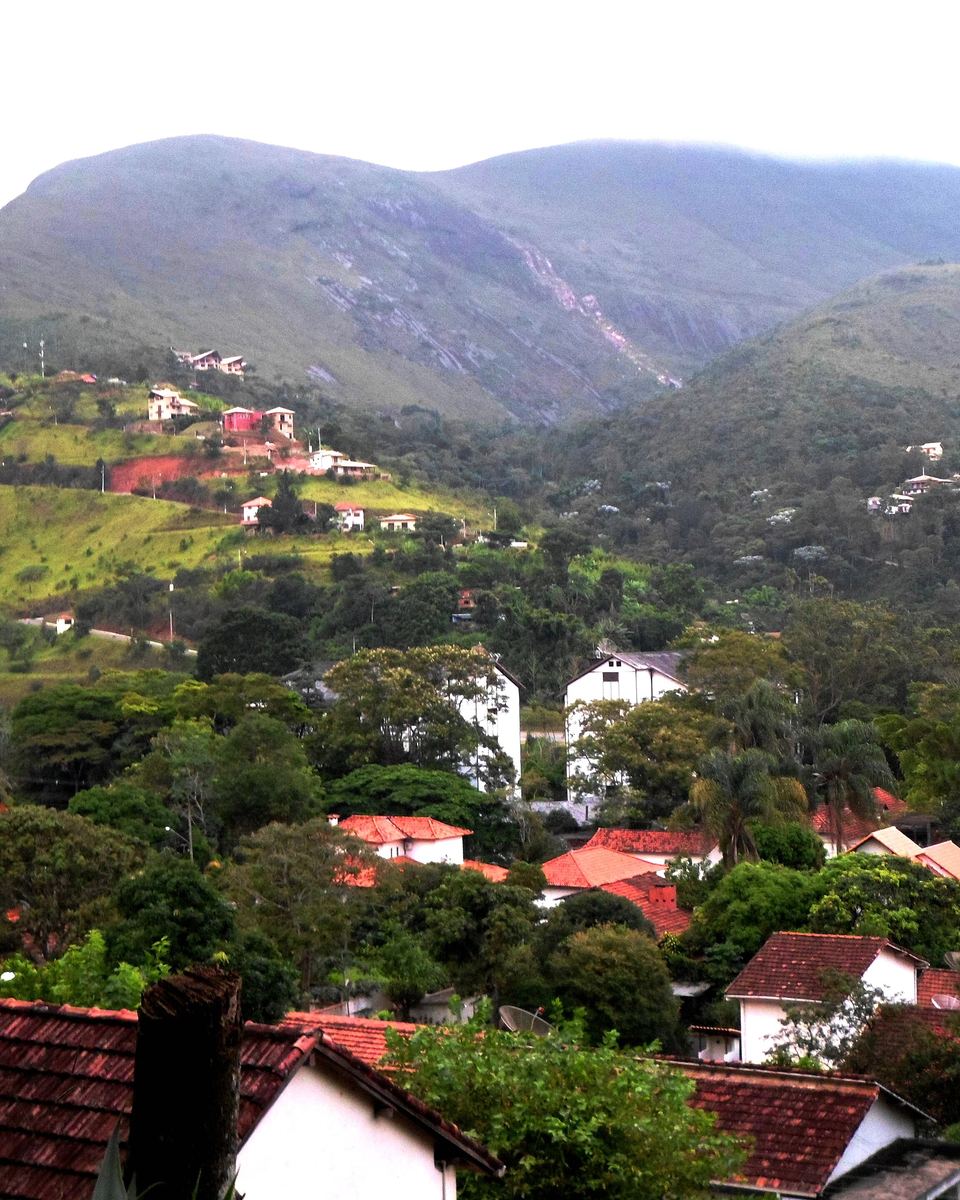 Vista de Itaipava, distrito de Petrópolis, com casas de telhado vermelho entre árvores e as montanhas verdes da serra ao fundo, num dia nublado.