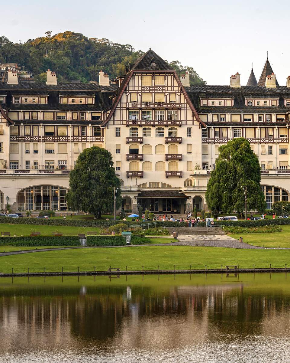 Palácio Quitandinha, em Petrópolis, prédio em estilo normando com telhados escuros, gramado e lago na frente e a mata da serra ao fundo.