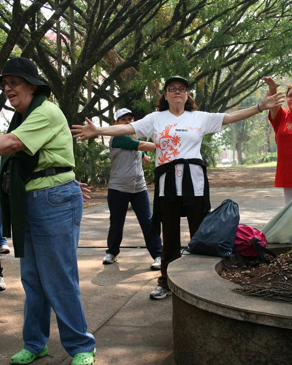 Grupo de adultos fazendo alongamento ao ar livre, com os braços abertos, numa calçada arborizada de Piracicaba.