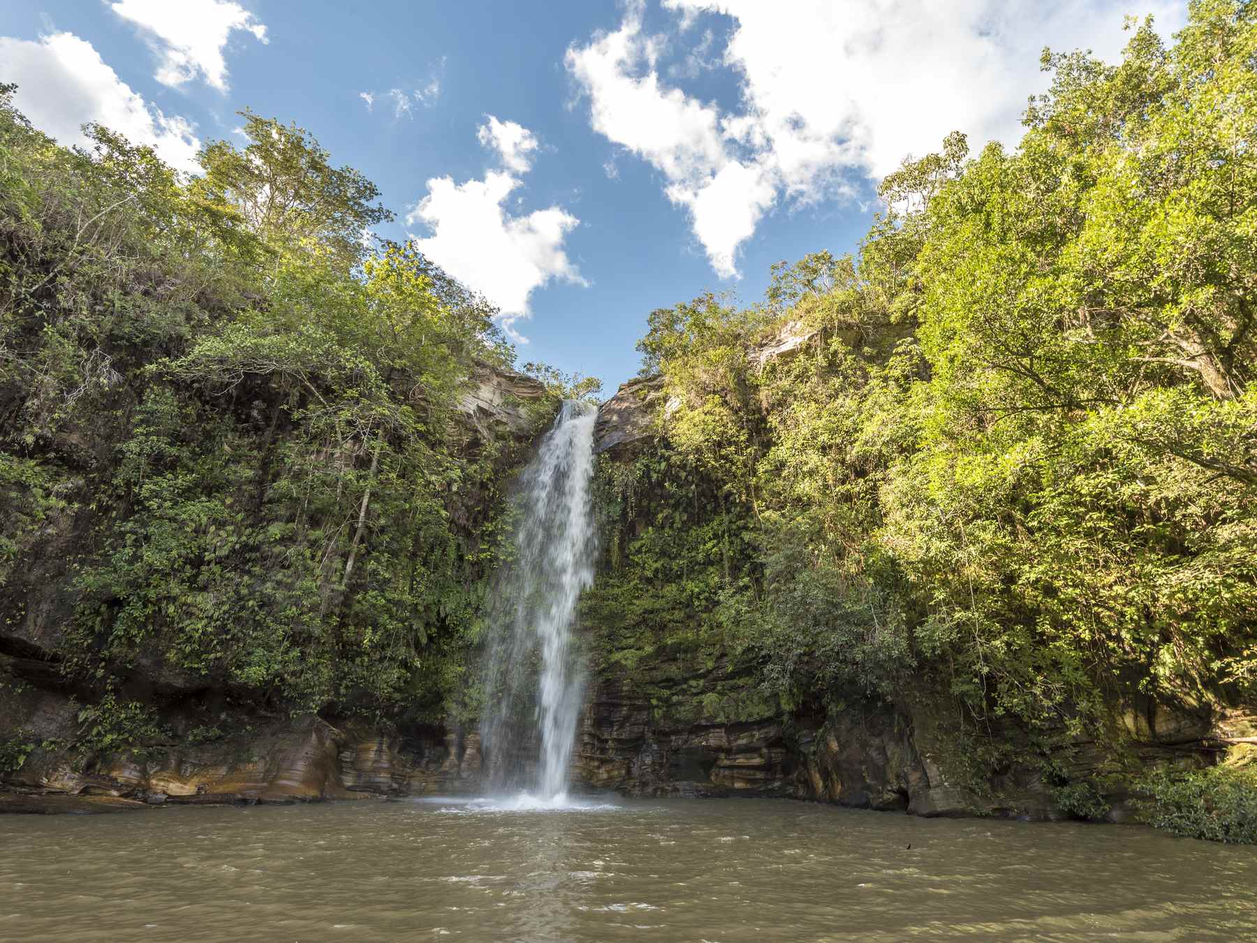 Cachoeira do Abade, em Pirenópolis, com a queda d'água fina descendo o paredão de pedra coberto de mata até um poço largo, sob céu azul com nuvens brancas.
