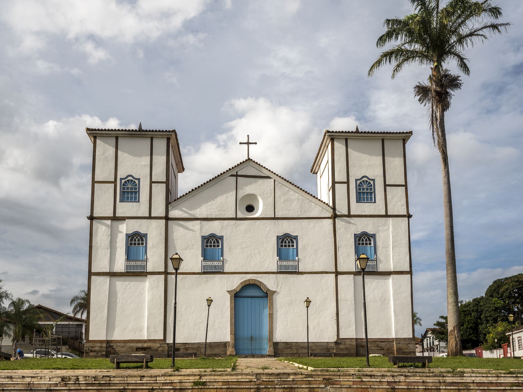 Fachada da Igreja Matriz de Nossa Senhora do Rosário, em Pirenópolis, branca com duas torres, portas azuis e uma palmeira alta ao lado, sob céu azul com nuvens.
