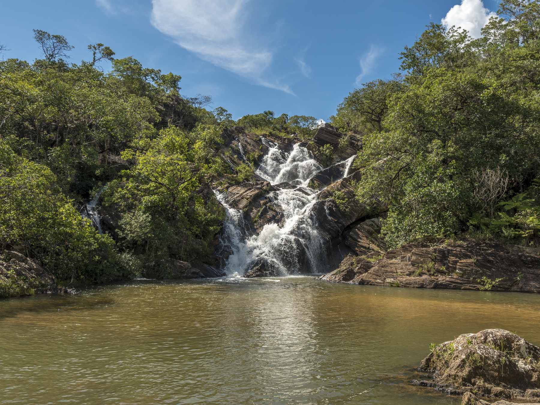 Cachoeira do Lázaro, em Pirenópolis, com a água branca descendo em degraus de pedra entre a mata até um poço largo de água cor de chá, sob céu azul.
