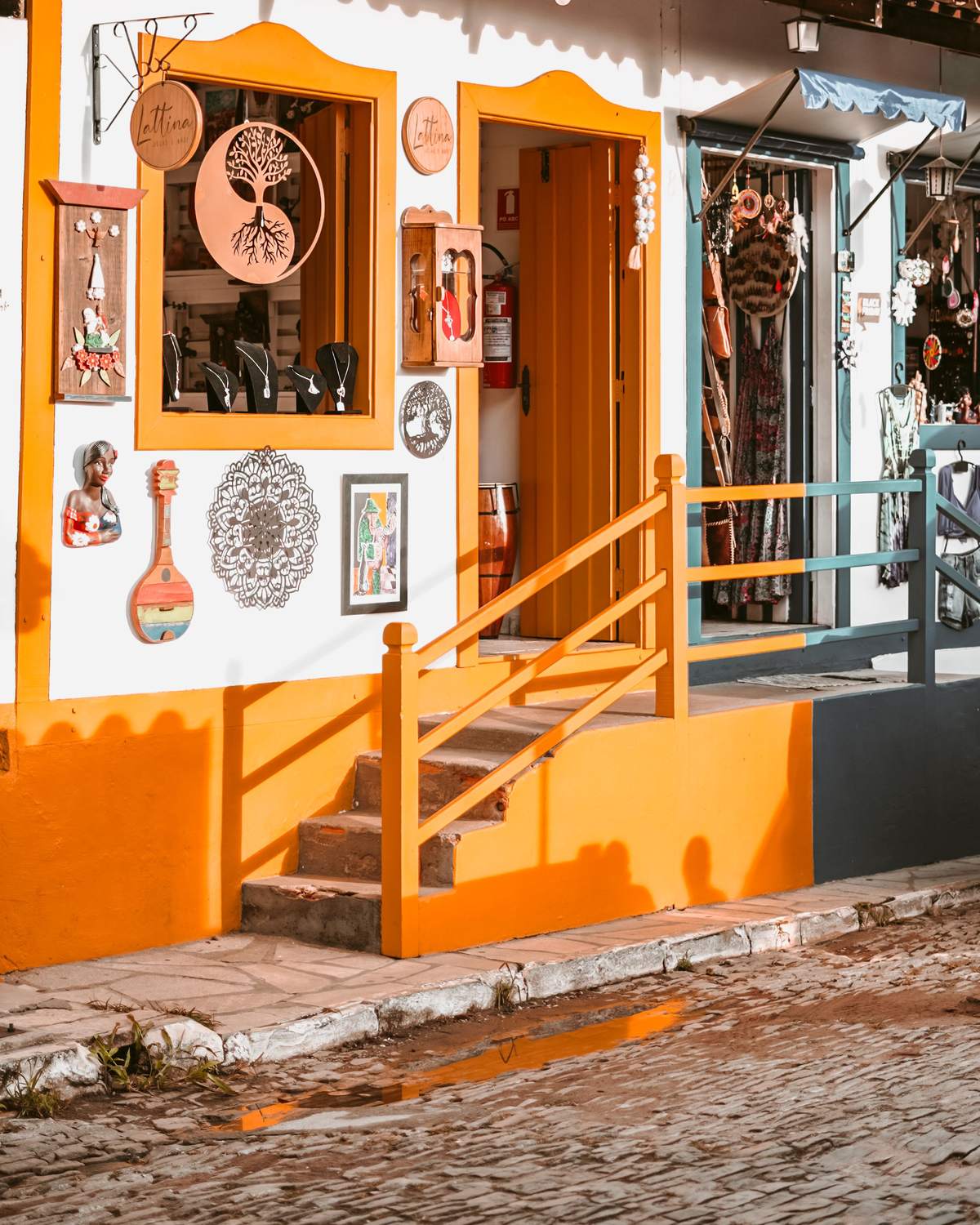 Fachada de uma loja de artesanato em Pirenópolis, pintada de branco e laranja, com mandalas de metal, quadros e peças penduradas na parede, porta aberta e escadinha de madeira sobre a rua de pedra.