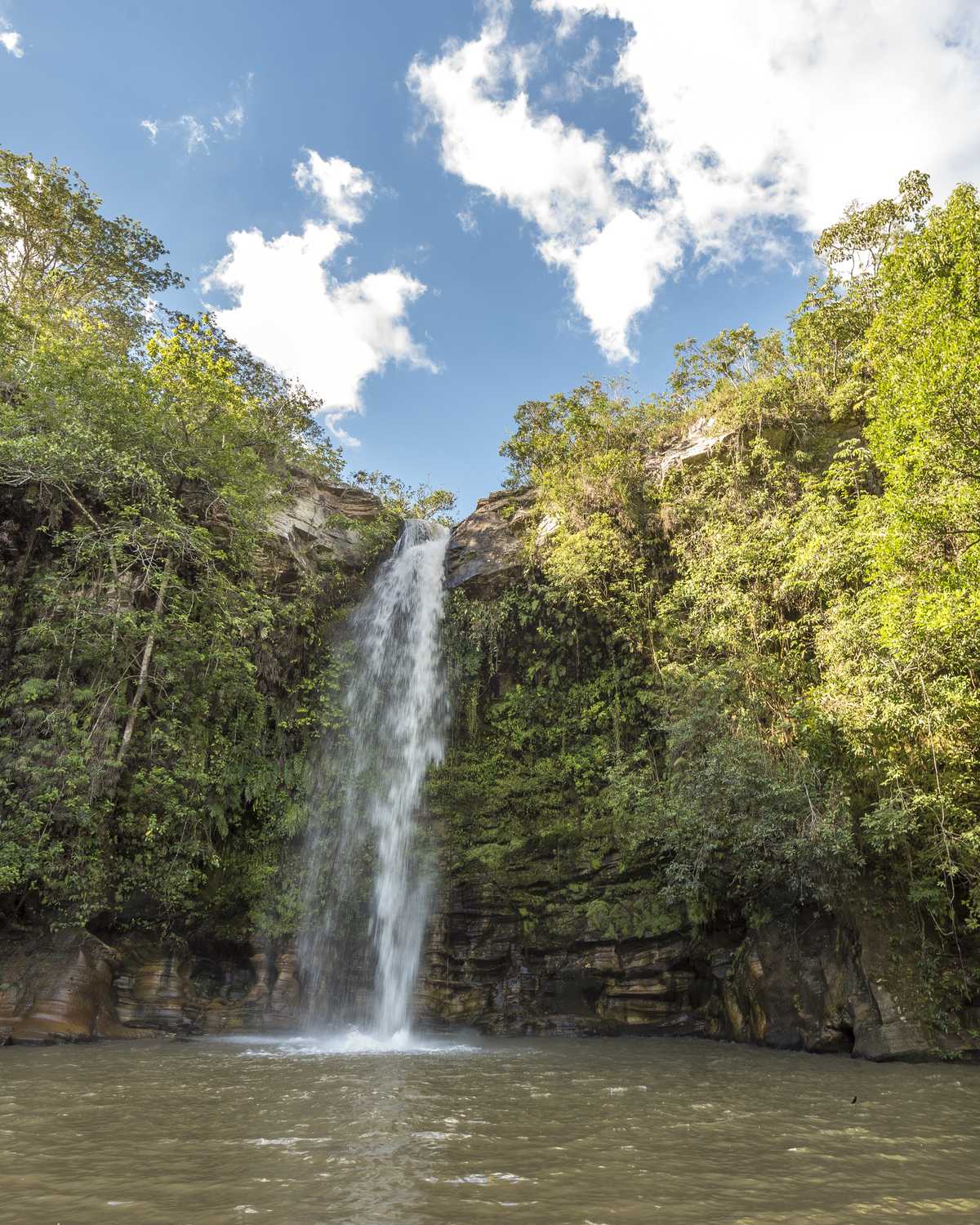 Cachoeira do Abade, em Pirenópolis, com a queda d'água fina descendo o paredão de pedra coberto de mata até um poço largo de água escura, sob céu azul com nuvens brancas.