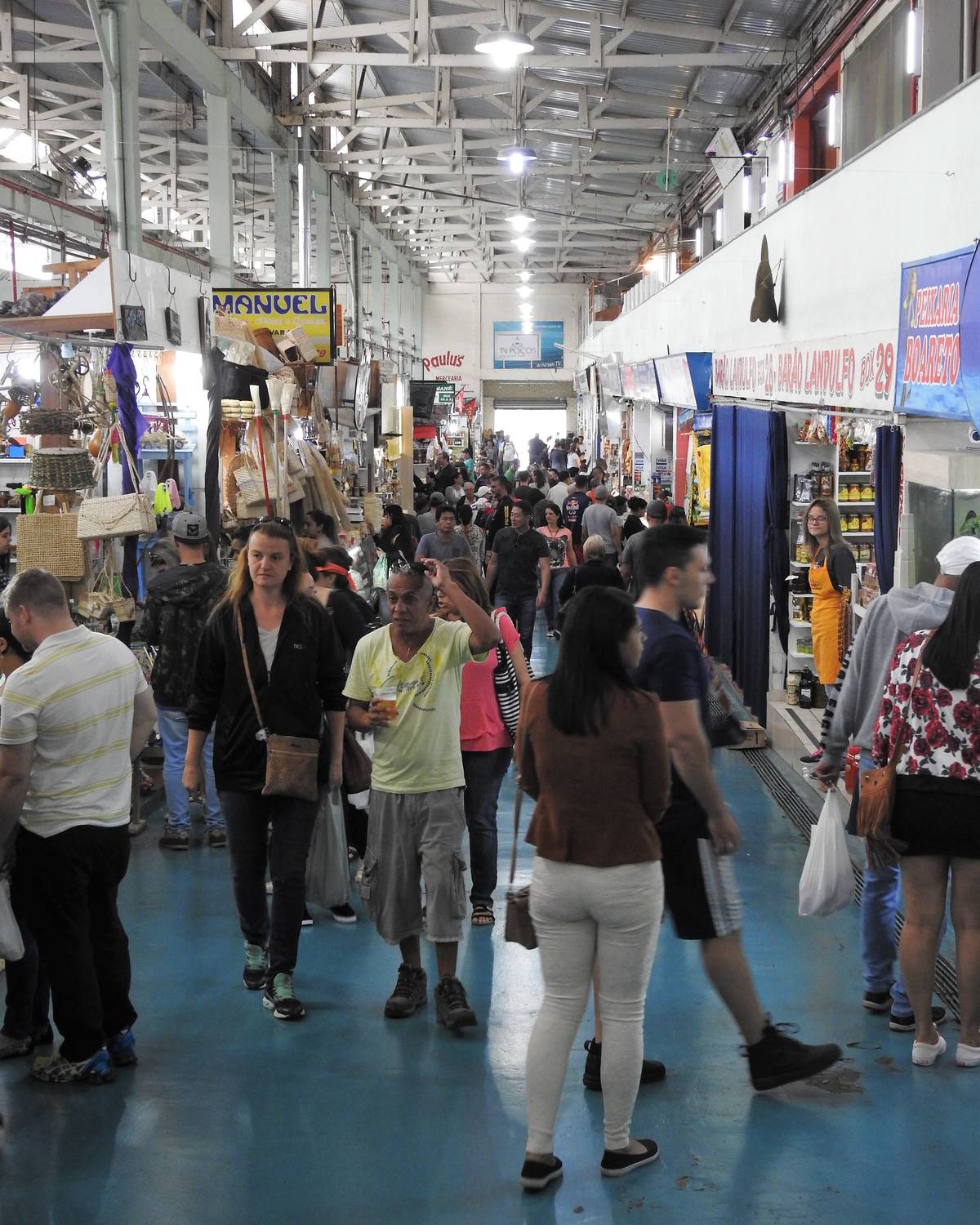Corredor do Mercado Municipal de Poços de Caldas cheio de gente, com bancas de cestaria e artesanato à esquerda, lojas à direita e clientes carregando sacolas.