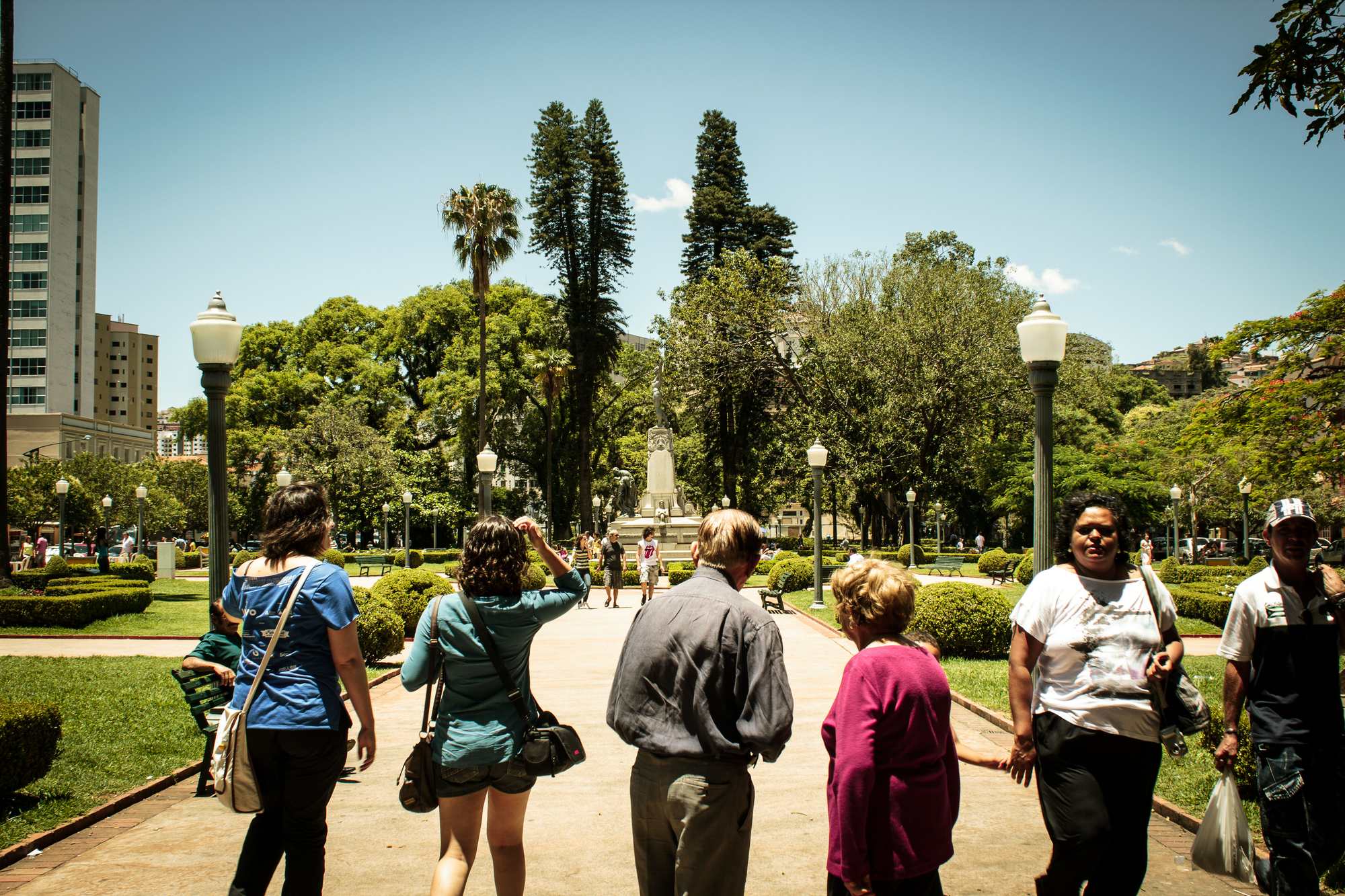 Moradores e turistas caminhando por uma alameda do Parque José Affonso Junqueira, no centro de Poços de Caldas, entre canteiros de buxinho, postes de ferro e araucárias, num dia de sol.