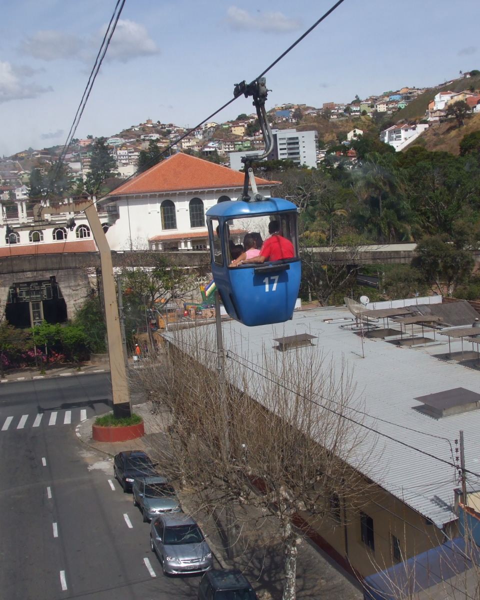 Cabine azul do teleférico de Poços de Caldas suspensa nos cabos, passando sobre uma rua do centro com carros estacionados, prédios baixos e o morro com casas ao fundo.