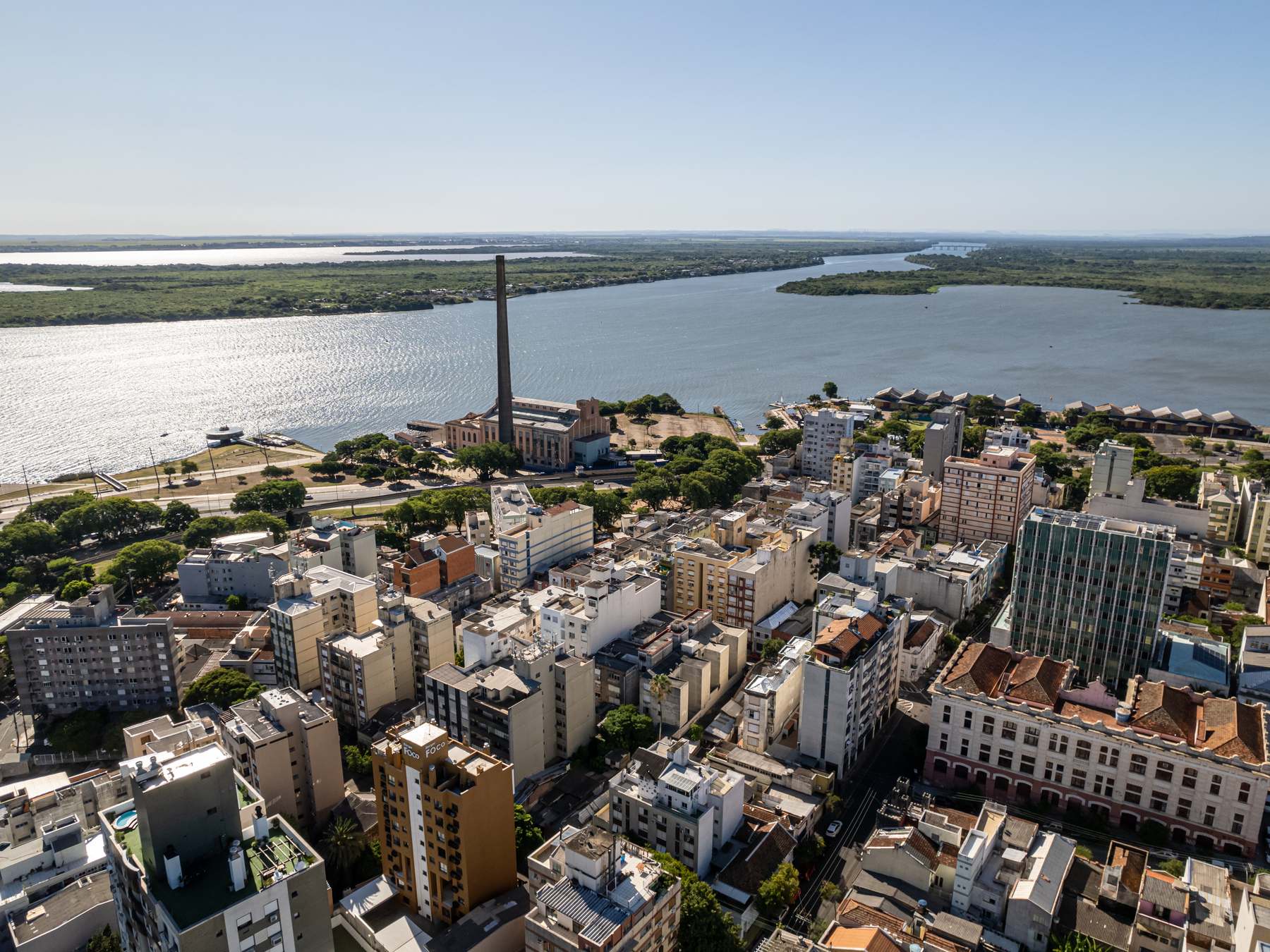 Vista aérea do Centro de Porto Alegre com a chaminé da Usina do Gasômetro, a orla e o Guaíba largo com as ilhas do delta do Jacuí ao fundo, num dia de sol.