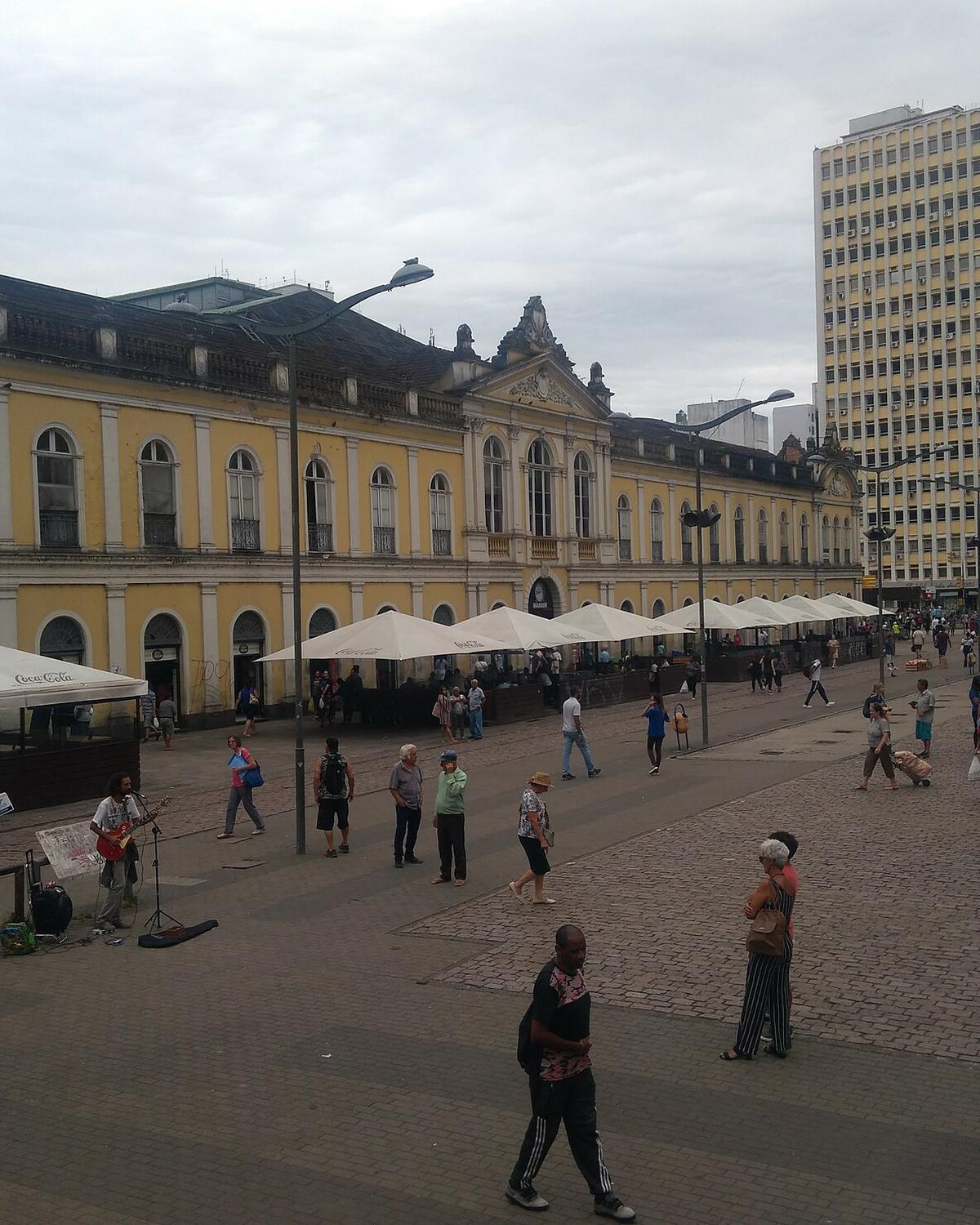 Fachada amarela do Mercado Público de Porto Alegre vista do Largo Glênio Peres, com ombrelones brancos, pedestres atravessando o largo e prédios altos do Centro ao fundo.