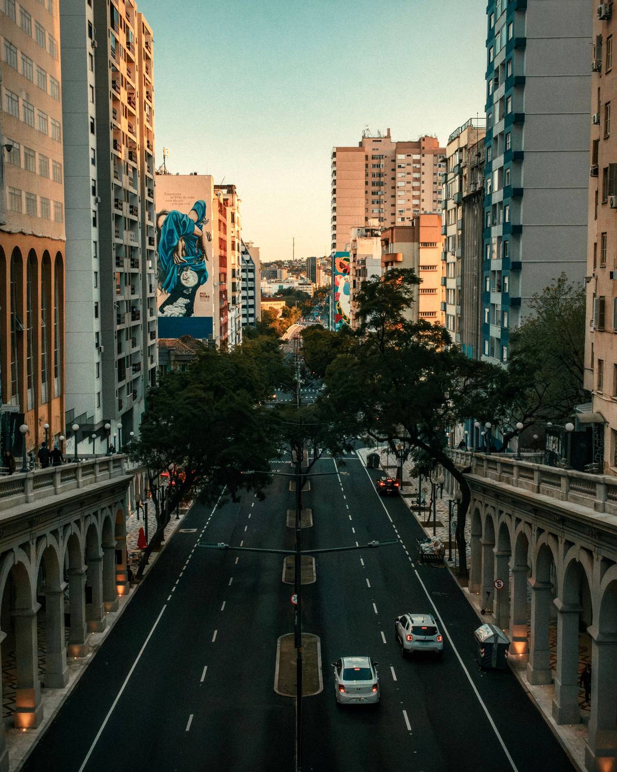 Avenida Borges de Medeiros vista do alto do Viaduto Otávio Rocha, em Porto Alegre, ao entardecer, com as arcadas do viaduto, prédios dos dois lados e um grande mural numa empena ao fundo.