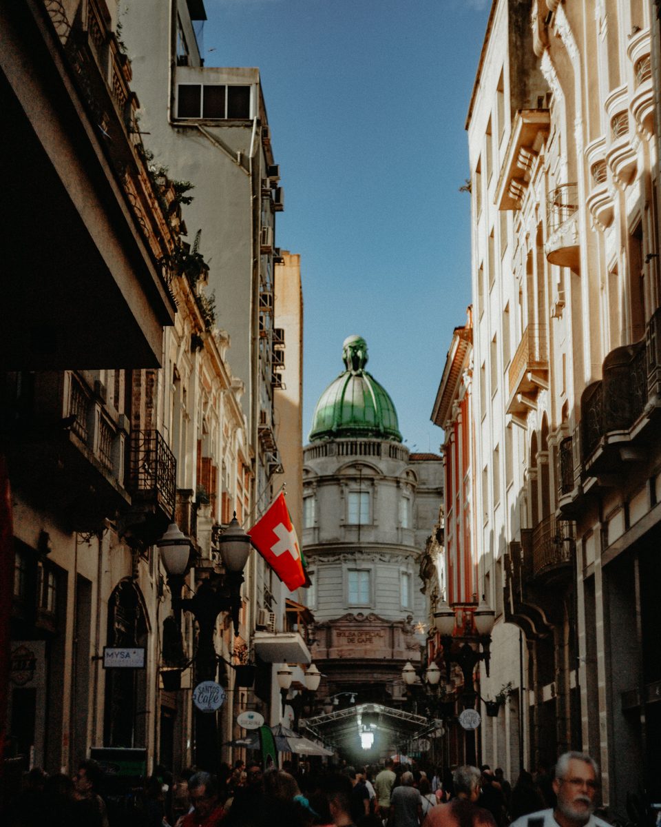 Pedestres numa rua estreita do Centro Histórico de Porto Alegre, entre prédios antigos de fachada trabalhada, sacadas de ferro e postes de luz, com uma cúpula verde ao fundo sob céu azul.