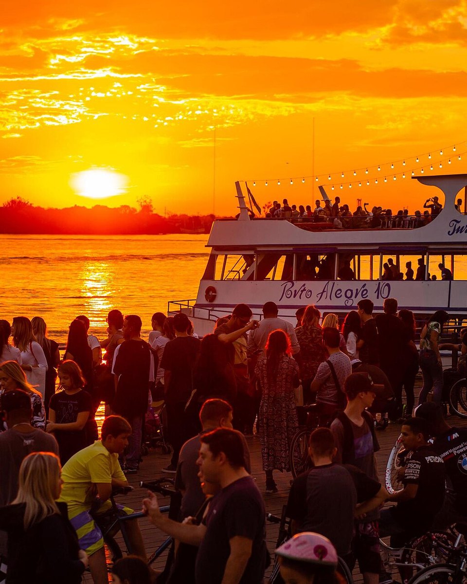 Multidão reunida na Orla Moacyr Scliar, em Porto Alegre, assistindo ao pôr do sol alaranjado sobre o Guaíba, com um barco de passeio e um veleiro na água.