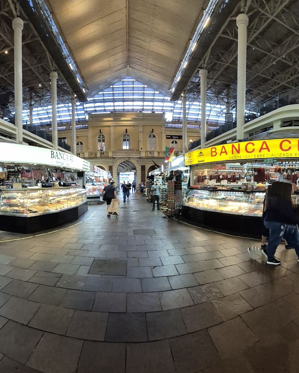 Interior do Mercado Público de Porto Alegre em vista panorâmica, com bancas iluminadas dos dois lados do corredor central, estrutura metálica no teto e pessoas caminhando pelo piso de pedra.