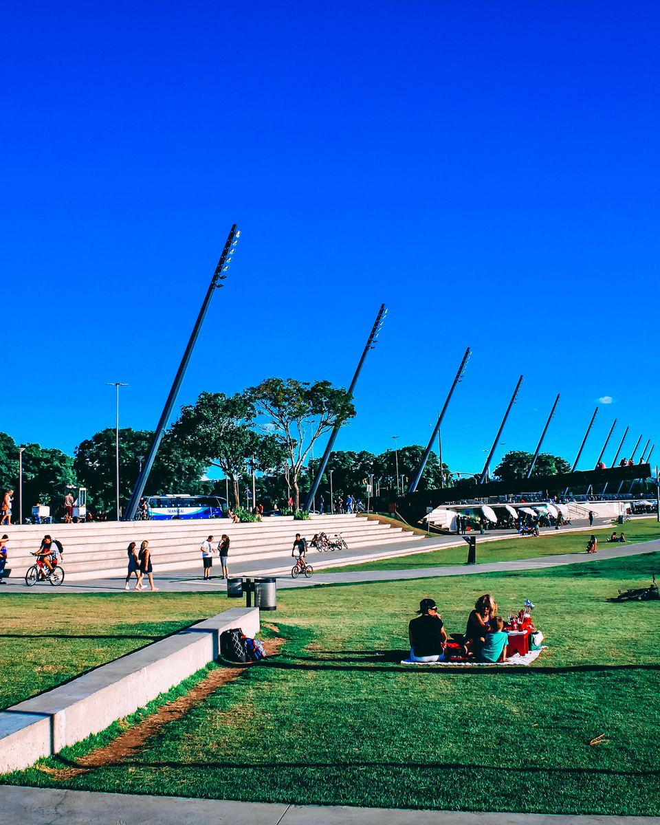 Pessoas sentadas e caminhando no gramado e na arquibancada da Orla Moacyr Scliar, em Porto Alegre, num dia de céu azul, com os postes altos inclinados da orla e o Guaíba ao fundo.