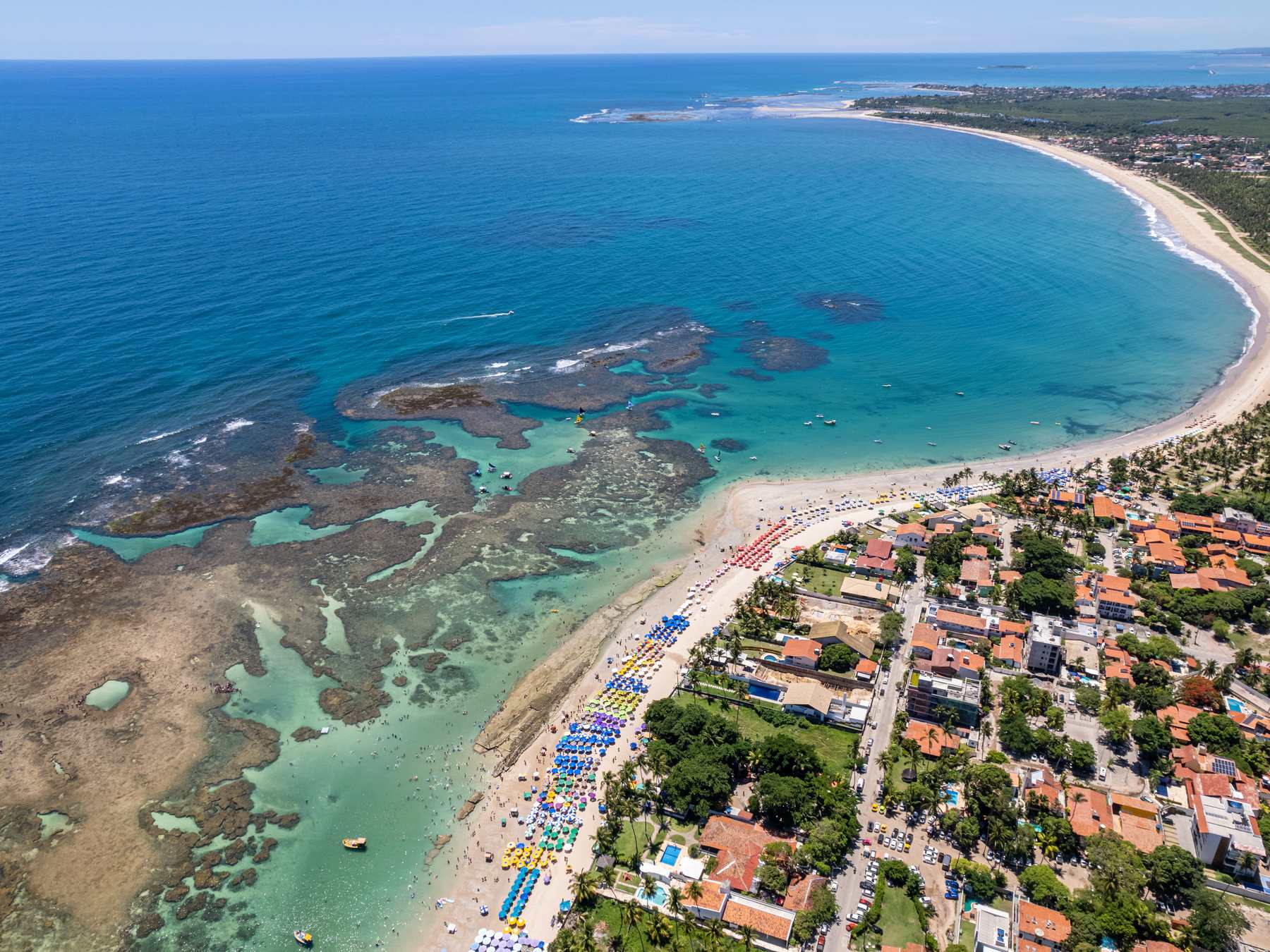 Vista aérea da orla de Porto de Galinhas, com a faixa de areia clara em curva, o mar azul-turquesa, os recifes escuros na água e a vila de casas e prédios baixos entre coqueiros.
