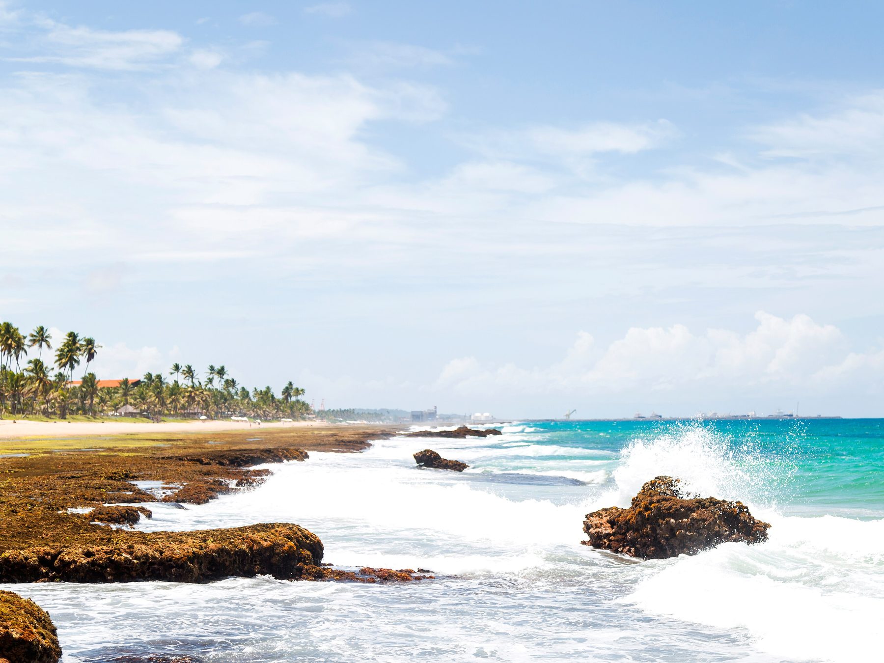 Praia de Muro Alto com as ondas quebrando em espuma branca sobre as pedras do recife, a água azul-turquesa e a linha de coqueiros ao fundo sob céu claro.
