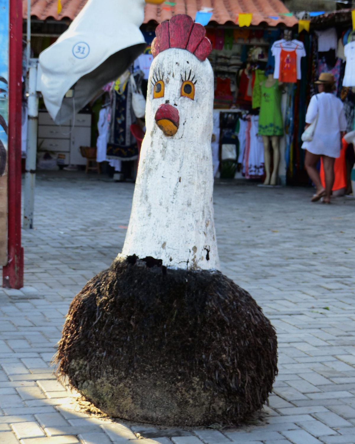 Escultura de galinha branca de crista vermelha, entalhada em madeira sobre a base de raiz de coqueiro, numa calçada de pedra da vila de Porto de Galinhas, com uma loja de roupas e turistas ao fundo.