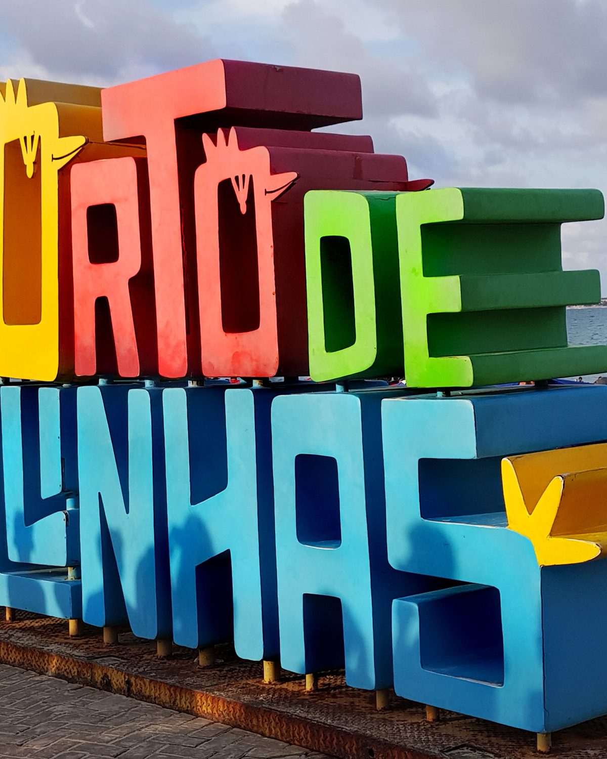 Letreiro colorido com as palavras Porto de Galinhas em letras grandes de vermelho, amarelo, verde e azul na orla, com turistas em volta e o mar ao fundo.