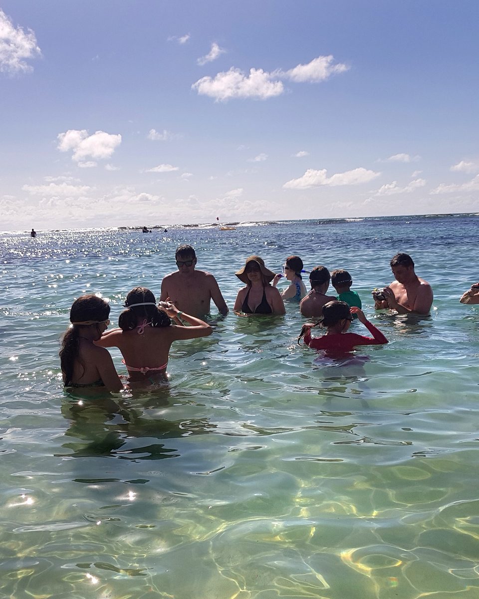 Turistas com água pela cintura nas piscinas naturais de Porto de Galinhas, na água verde-clara e transparente, sob céu azul com poucas nuvens.