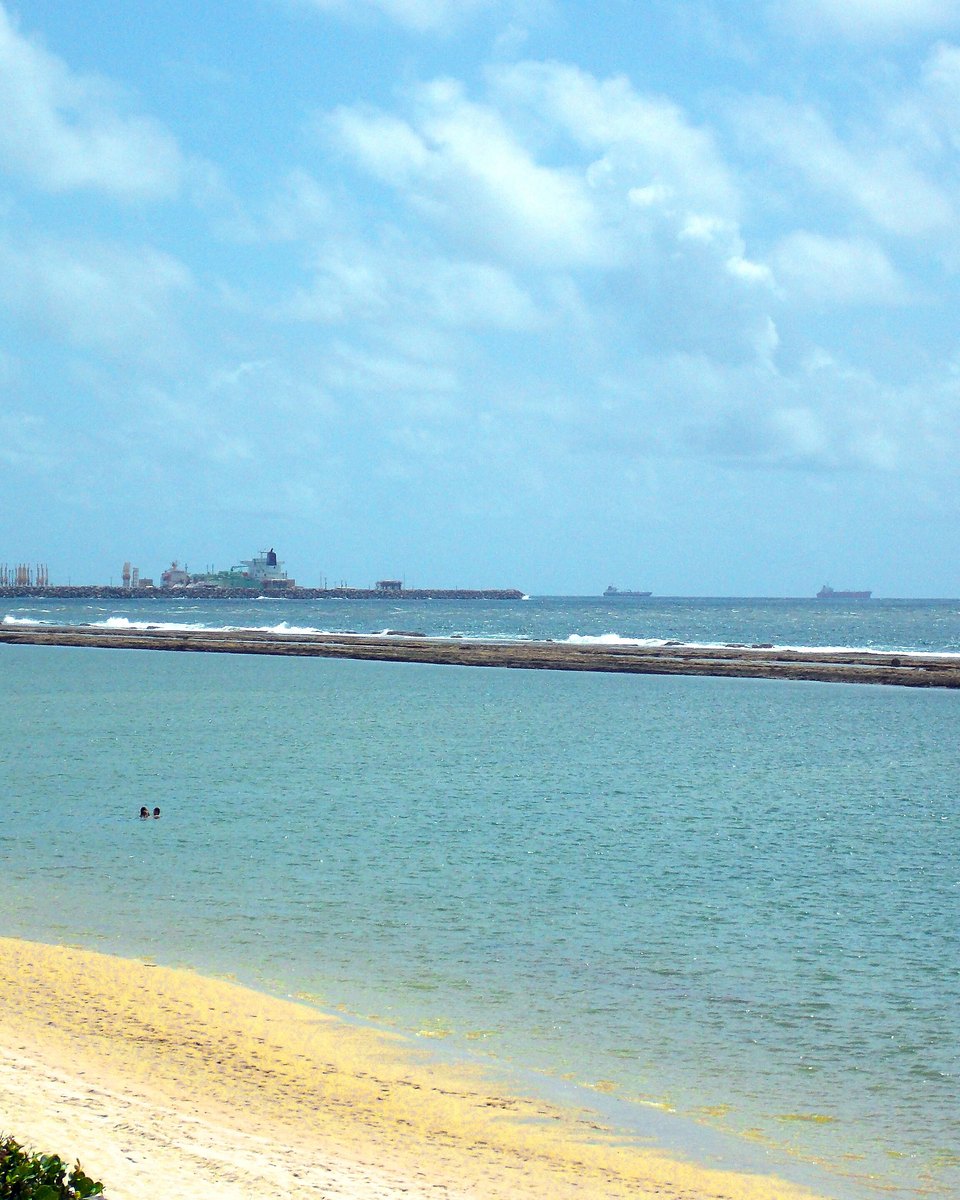 Mar calmo e azul-claro visto do alto de uma encosta em Muro Alto, com a faixa de areia clara em curva e, no horizonte, os molhes, os guindastes e os navios do Porto de Suape.