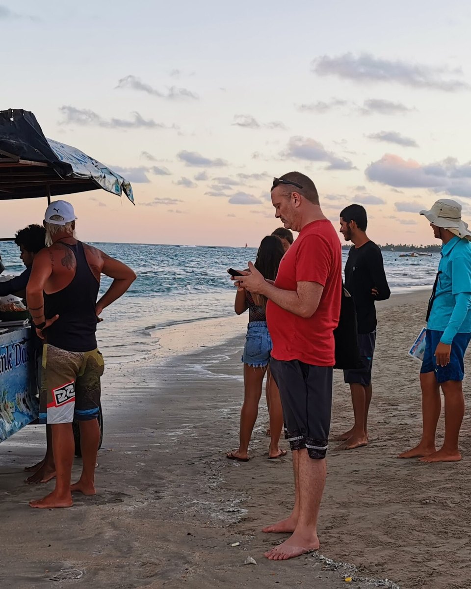 Carrinho de drinques com toldo colorido na beira do mar em Porto de Galinhas, com o vendedor atendendo e turistas conversando na areia no fim da tarde.