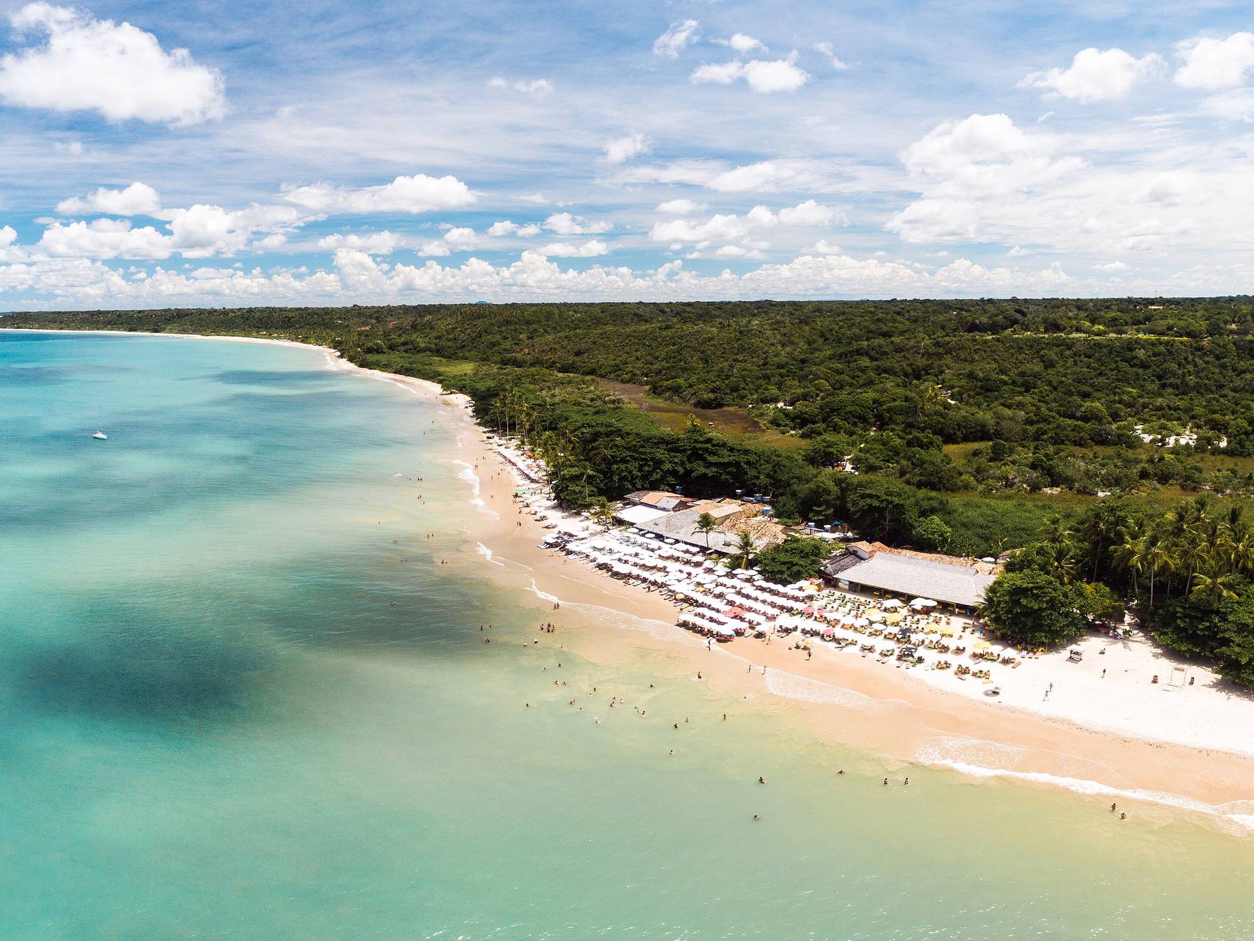 Vista aérea da Praia dos Coqueiros, em Trancoso, com o mar raso azul-turquesa, a areia clara com guarda-sóis e a mata atlântica ao fundo.