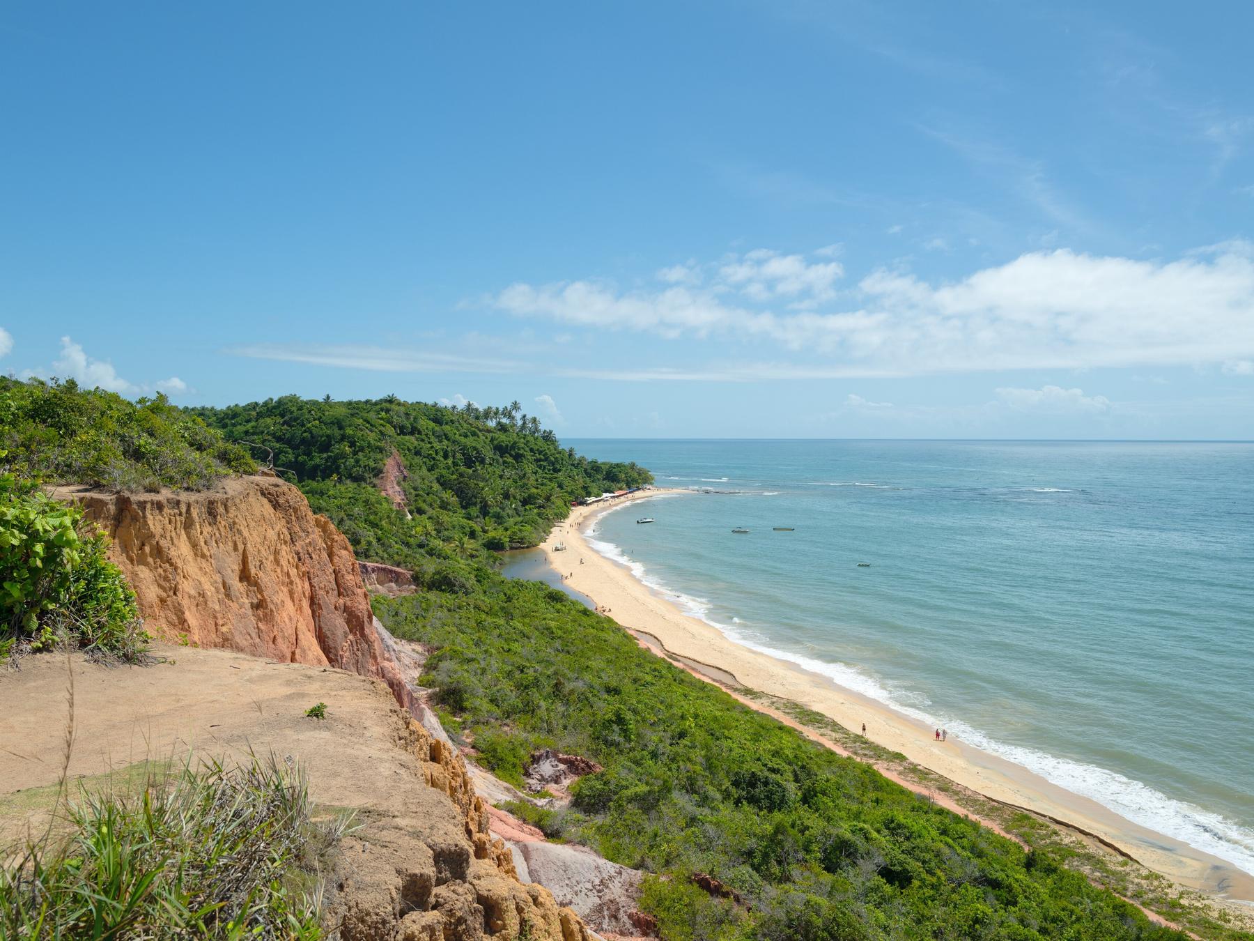 Falésias alaranjadas cobertas de mata vistas do alto em Arraial d'Ajuda, com a praia comprida e o mar azul sob o céu com nuvens.