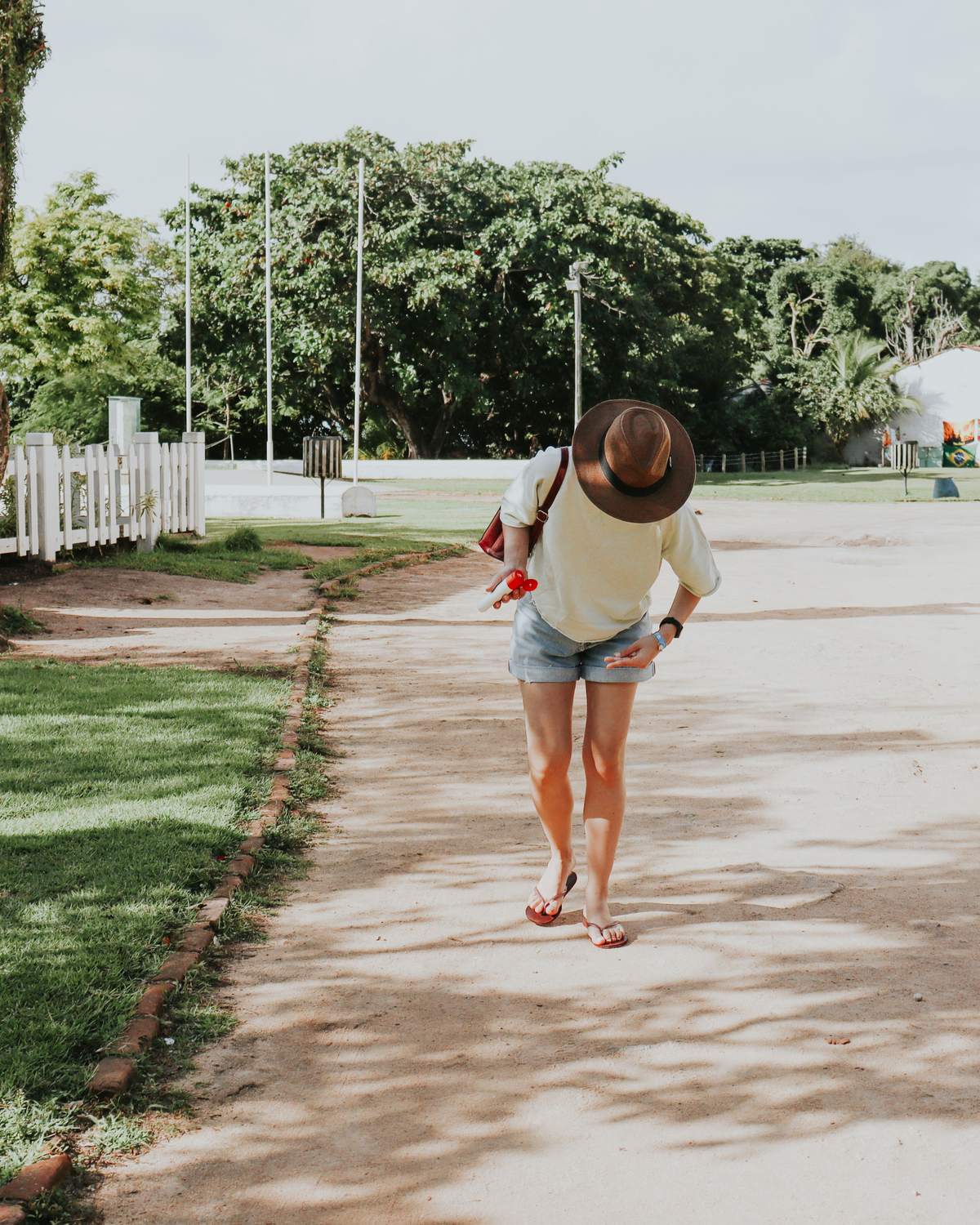 Mulher de chapéu, camiseta clara e short jeans caminhando de chinelo por uma rua de terra da Cidade Histórica de Porto Seguro, entre uma casa branca de janelas vermelhas e o gramado.
