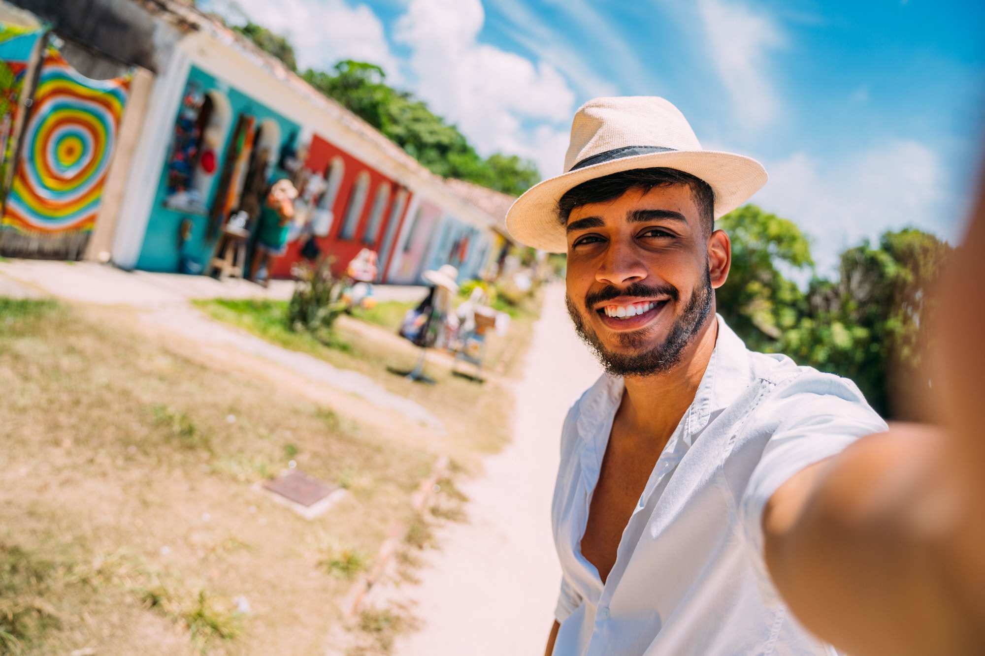 Turista de chapéu de palha e camisa branca sorrindo e fazendo selfie com o celular numa rua de terra da Cidade Histórica de Porto Seguro, com a fileira de lojas de artesanato coloridas ao fundo.