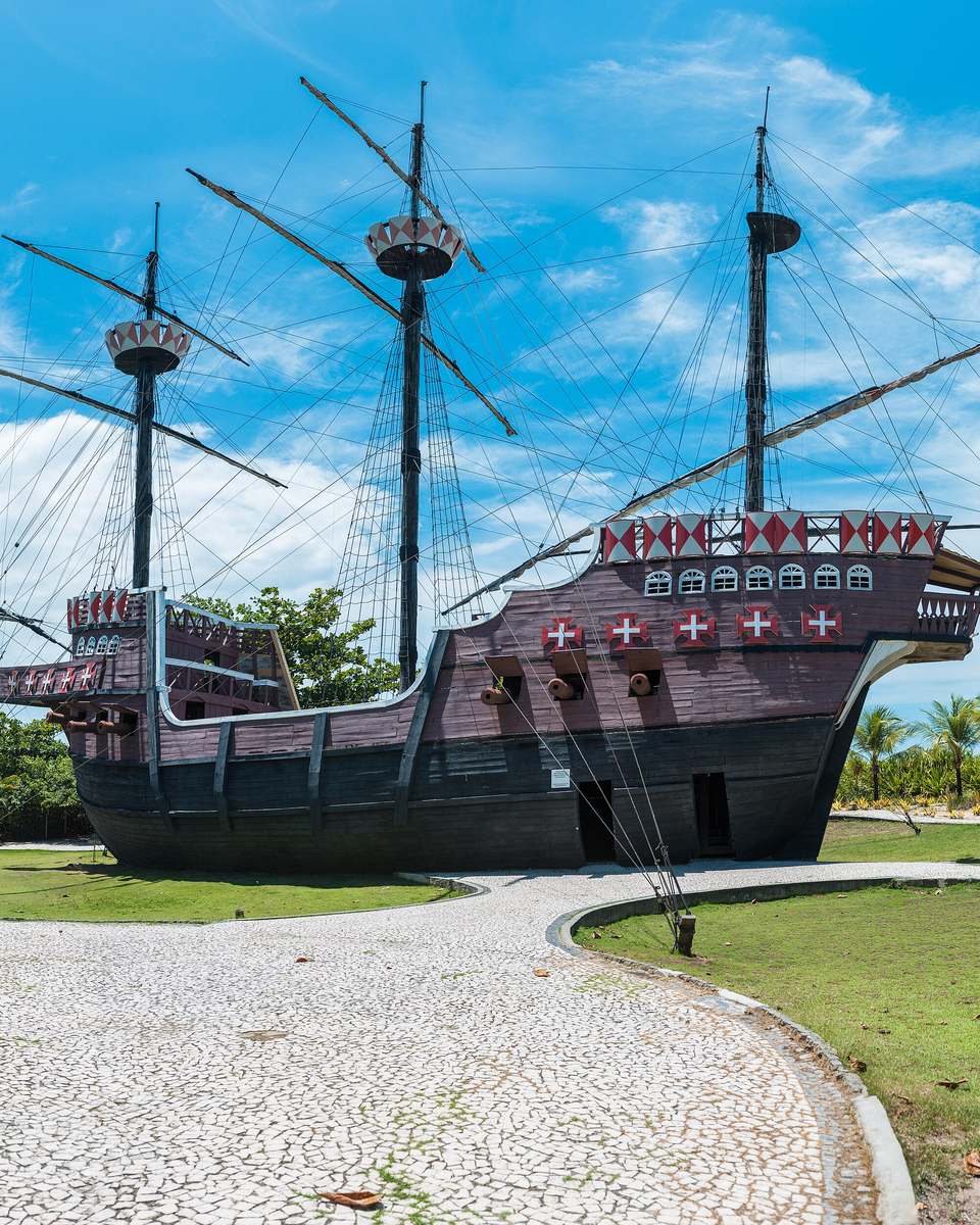 Réplica em tamanho real de uma nau portuguesa de três mastros com velas recolhidas no Memorial da Epopeia do Descobrimento, em Porto Seguro, sobre o gramado, com coqueiros e o céu azul ao fundo.