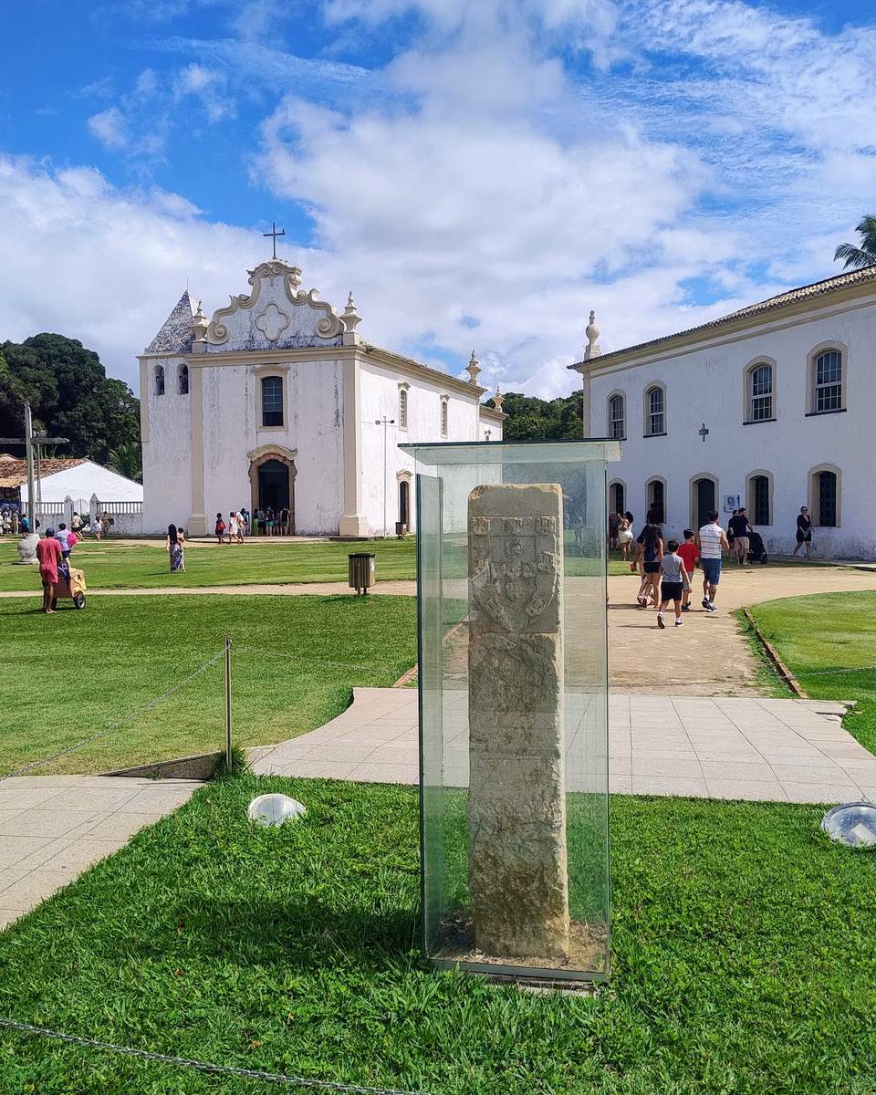 Cidade Alta de Porto Seguro num dia de sol, com a igreja e o casario coloniais brancos, visitantes caminhando pelo gramado e o Marco do Descobrimento protegido por uma caixa de vidro em primeiro plano.
