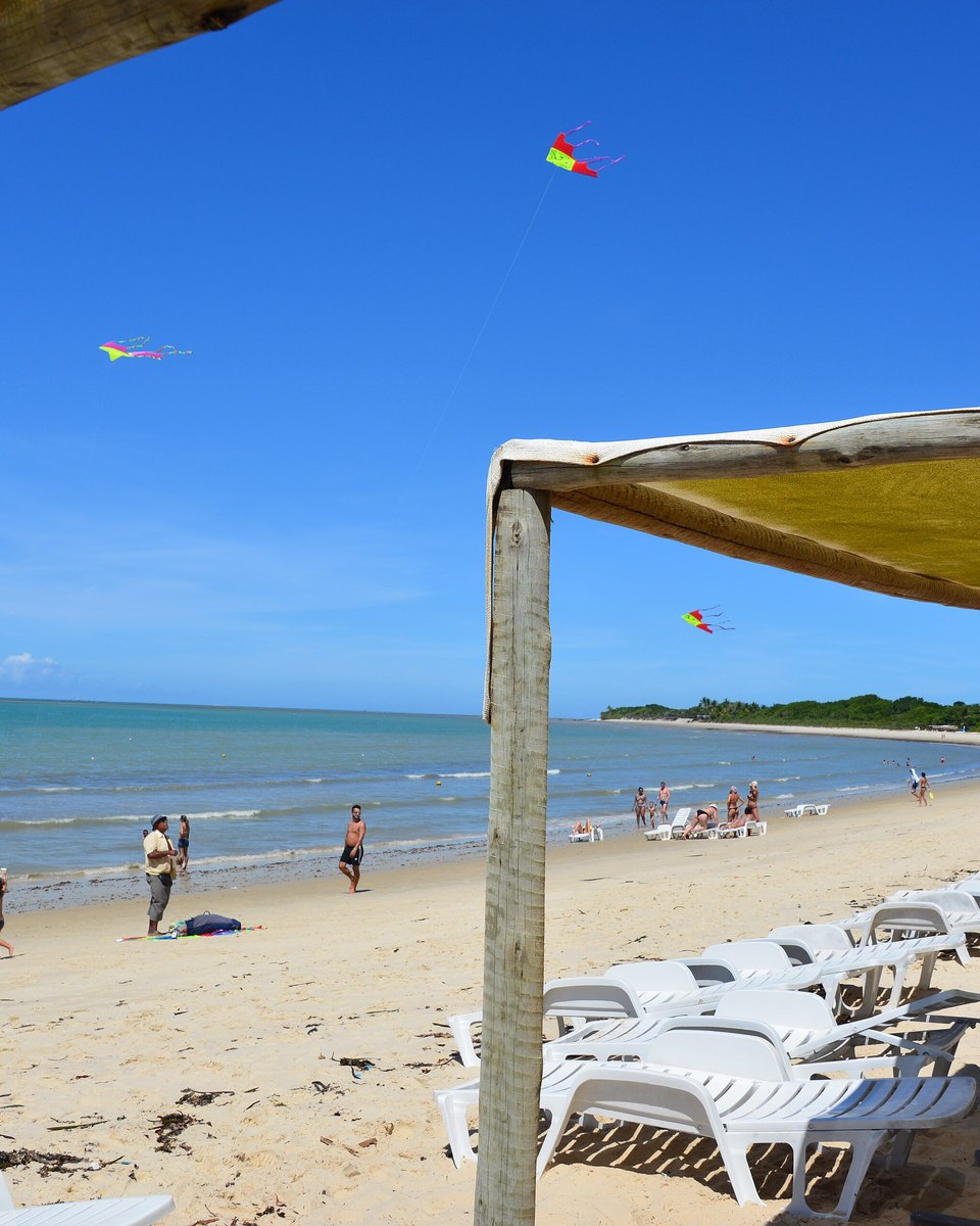 Praia de Porto Seguro vista de baixo de uma barraca de lona, com espreguiçadeiras brancas na areia, banhistas na beira do mar calmo e três pipas coloridas no céu azul.